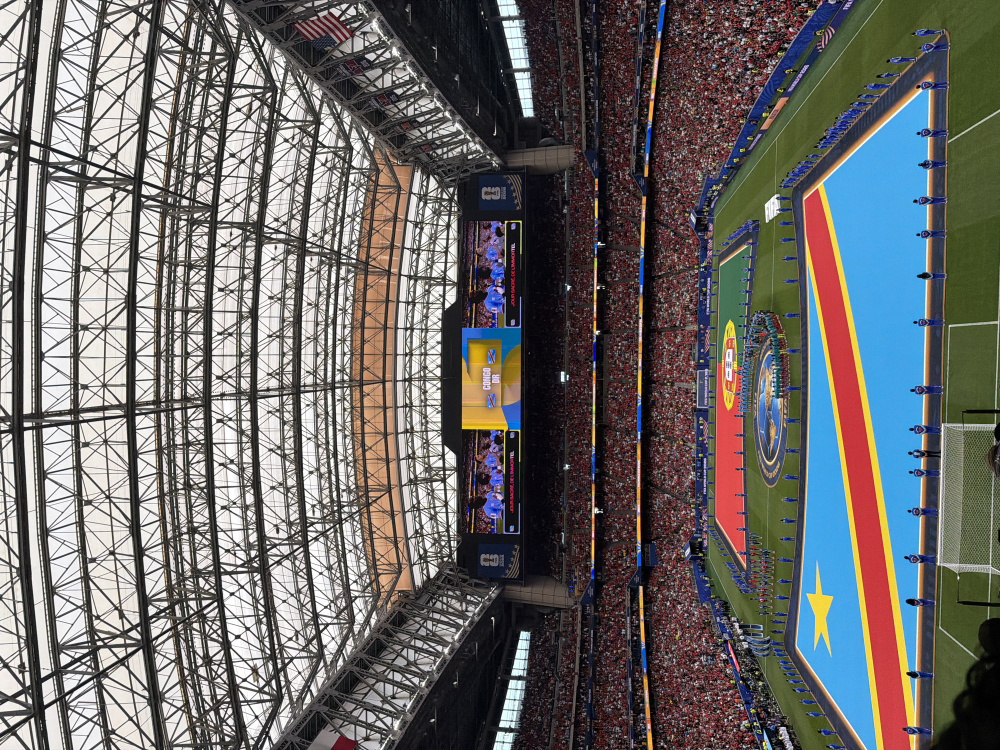 The image captures a large-scale event taking place in a stadium, likely during the daytime, as evidenced by the bright natural light filtering through the stadium's structure. The scene is dominated by a vast crowd of spectators filling the stands, wearing predominantly red attire, creating a sea of color. In the center of the frame, a soccer field is visible with two distinct flags displayed prominently on the grass. One flag is the Democratic Republic of Congo's flag, characterized by its sky blue field, a red stripe bordered by yellow, and a yellow star. Adjacent to it is another flag, which appears to be Portugal's, featuring green and red. Players, dressed in blue uniforms, are lined up on the field, suggesting a pre-game ceremony or the start of a match between the two nations. A large screen at one end of the field displays images of players and text. Text visible on the screen includes "CONGO," "D.R.," and "JOUR SACRÉ DE L'IMMORTEL." Another screen shows a logo with "25" and "STARS". The architectural elements of the stadium, with its intricate metal framework and translucent roof, are also a significant part of the scene. The atmosphere seems charged with anticipation and excitement, typical of a major sporting event. The presence of American flags at the top of the stadium structure indicates the event is being held in the United States, specifically Houston, as suggested by the location context.