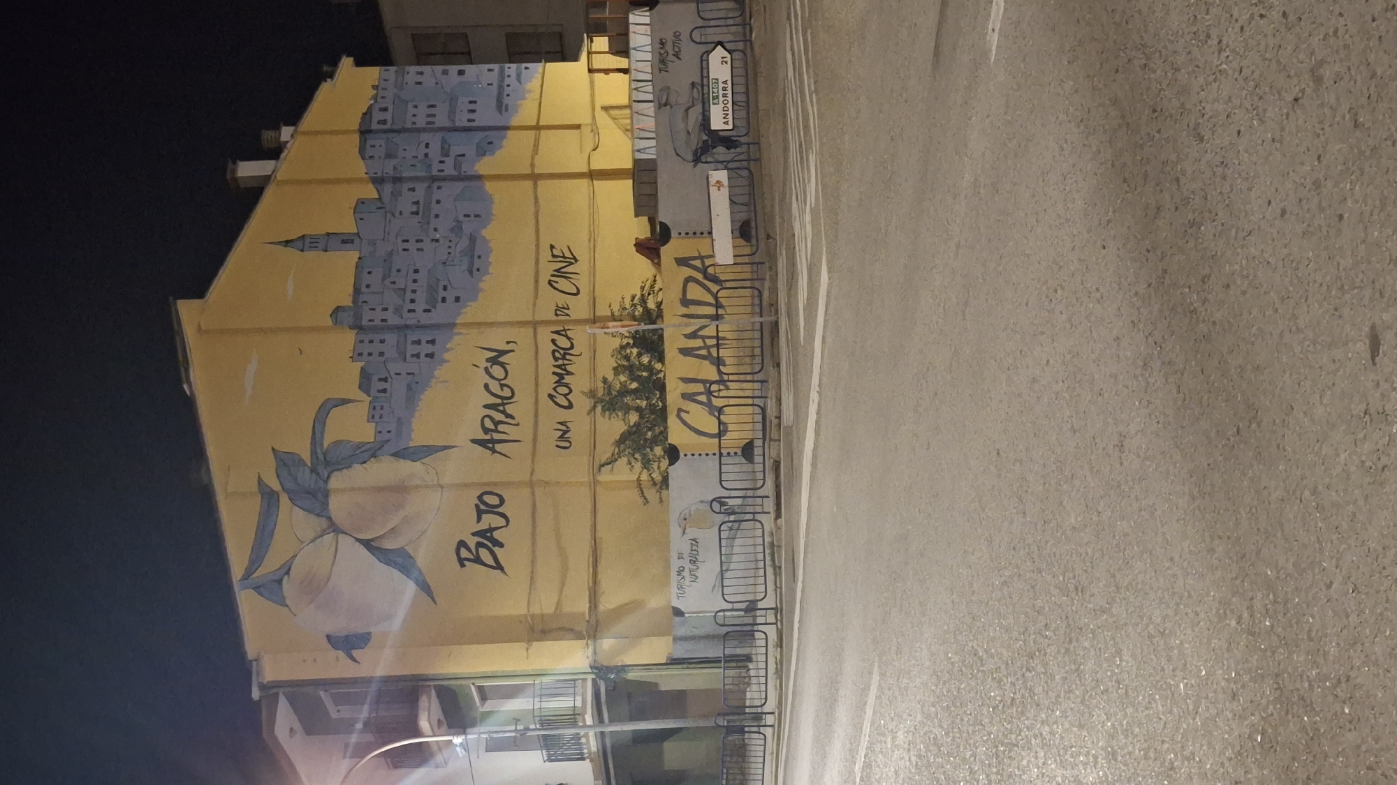 The image shows a mural painted on the side of a building in Calanda, Spain. The mural depicts a city skyline and a branch with two peaches. The text on the mural reads "BAJO ARAGÓN, UNA COMARCA DE CINE" and "CALANDA". There is also a drawing of a bird with the text "TURISMO Y NATURALEZA" below it, and another drawing of a figure cycling with the text "TURISMO ACTIVO" and "A-1407 ANDORRA 21". The mural is illuminated by streetlights. The foreground shows a street with a pedestrian crossing and a metal fence. The building is located in a town, likely in an urban area given the street and fence. The overall impression is a decorative mural advertising tourism in the region.