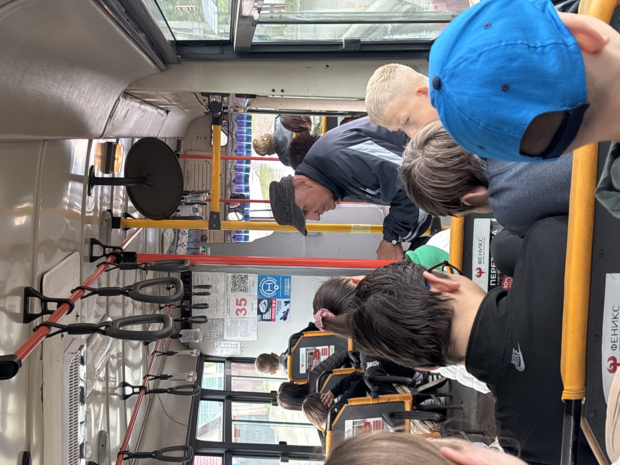 The image captures the interior of a public transport vehicle, likely a bus or trolleybus, during daytime. The scene is observed from a slightly elevated perspective, looking towards the front of the vehicle.
In the foreground, the back of a young person wearing a bright blue baseball cap is visible, alongside another individual with dark hair. In the mid-ground, an older man wearing a dark cap and jacket stands, looking forward or down. Next to him is a young boy with blonde hair. Further back, several other passengers, mostly appearing to be children or young adults, are seated or standing, seen primarily from behind.
The interior features numerous yellow vertical poles and red horizontal handrails equipped with dark grey grab handles for standing passengers. Seats with distinctive yellow frames are partially visible towards the right. A circular mirror or fan-like device is mounted on a pole near the ceiling on the left side. Windows offer glimpses of the outside, revealing buildings and possibly trees, indicating it is daytime.
Various posters and informational signs are affixed to the vehicle's walls. Visible text includes "ФЕНИКС" (Phoenix), which appears to be a company name, "35" (possibly a route number), and "АРЕНДА ТРАНСПОРТНЫХ БОЛЬНЫХ" (Rent of Transport for Sick/Patients), indicating a specialized transport service. A "NIKE" logo is visible on a black jacket worn by one of the passengers. Other fragmented text includes "ПЕРЕТ", "и рестар", and "-10%", likely part of an advertisement for a service or discount from the "Phoenix" company. The overall atmosphere suggests a routine journey or commute.