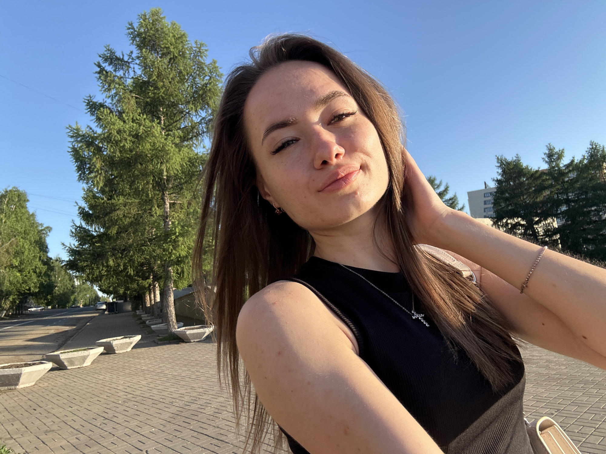 A young woman is featured in a close-up outdoor selfie, captured from a low angle, looking upwards towards a clear blue sky. She has long brown hair, pulled back slightly by her left hand which rests near her ear. She is wearing a black sleeveless top and a silver-colored necklace with a small cross pendant. A bracelet is visible on her right wrist, and a light-colored bag or strap is partially seen on her right side. Her expression is a relaxed smirk or slight smile, with her face illuminated by warm, directional sunlight, highlighting freckles on her nose and cheeks.
The background depicts a sunny urban or suburban street scene, suggesting late afternoon or early evening. Tall green coniferous trees line a paved sidewalk with several concrete planters arranged in a row. A road with a few parked cars is visible further down the street to the left. In the distance, on the right, generic buildings are nestled among more trees, all under a bright, cloudless blue sky, indicating clear and pleasant weather. There is no visible text in the image.