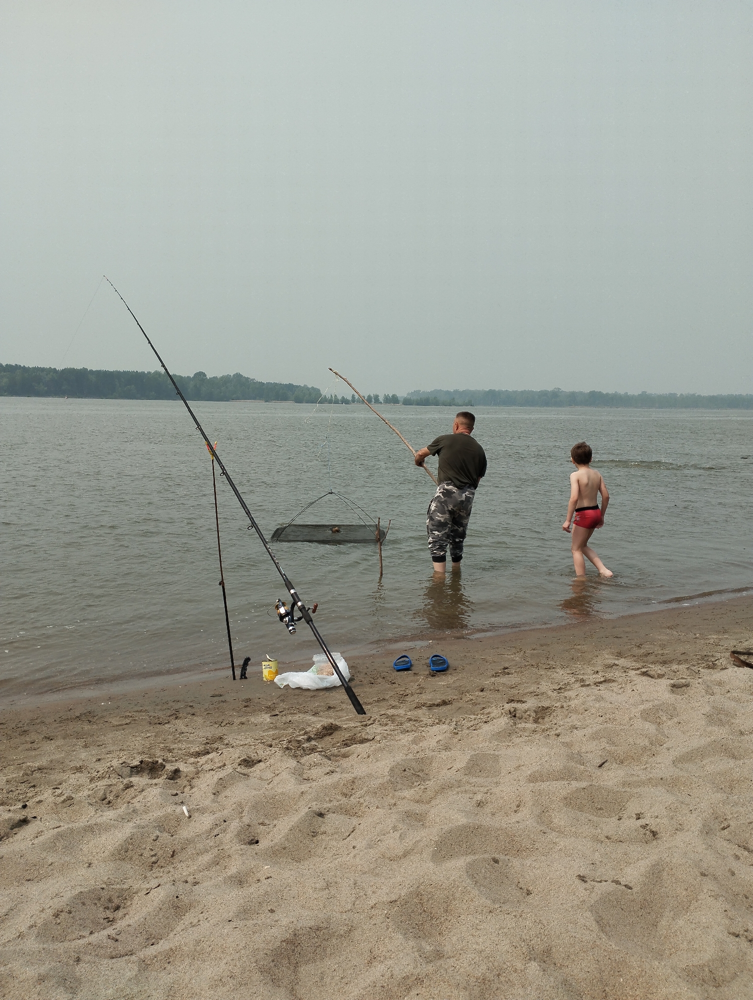 In Bibikha, Russia, on a sandy shore bordering a large body of water under an overcast sky, a man and a young boy are fishing. The man, wearing a dark green t-shirt and camouflage pants, stands in the water and holds a fishing rod, appearing to be casting or retrieving. The boy, in red swim trunks, walks in the shallow water nearby, facing away from the viewer.
Several fishing rods are set up on the sand near the water's edge. One prominent rod is upright, with its reel visible. Next to it are a can of what appears to be food, a plastic bag, and a smaller, thinner fishing rod. In the water, a net-like structure, possibly for catching fish, is partially submerged. Two blue flip-flops lie on the sand in the foreground. The overall atmosphere suggests a leisurely, familial outing by the water. The time of day appears to be daytime, indicated by the general lighting conditions, and the weather is hazy or foggy, obscuring the distant tree line.