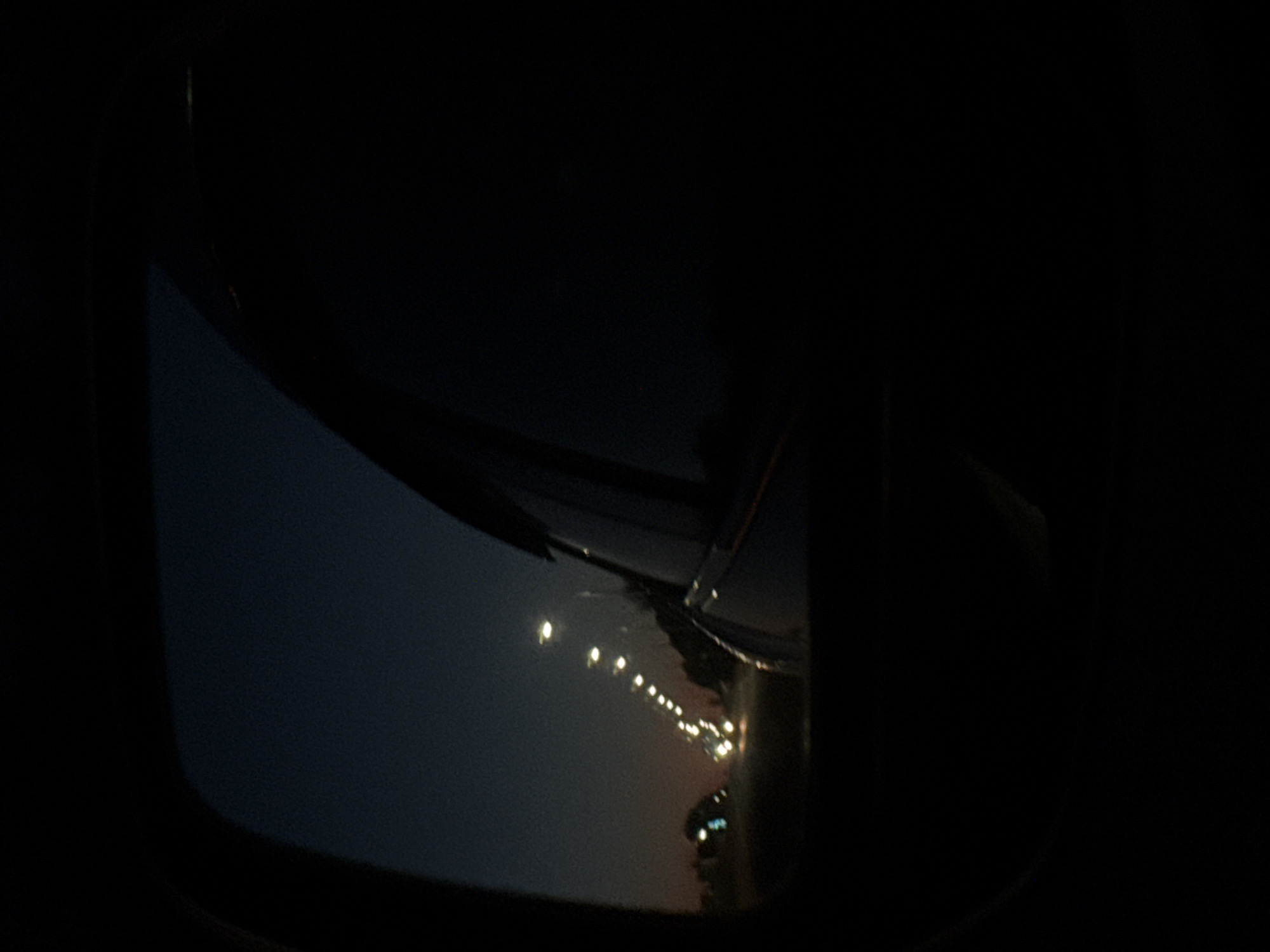 The image shows a view from an airplane window at night. The wing of the plane is partially visible in the frame, reflecting lights from the ground. Below the wing, a series of lights suggest a runway or city lights at night. The darkness surrounding the window indicates it is night, and only illuminated objects are visible. Location: Unknown, Unknown.