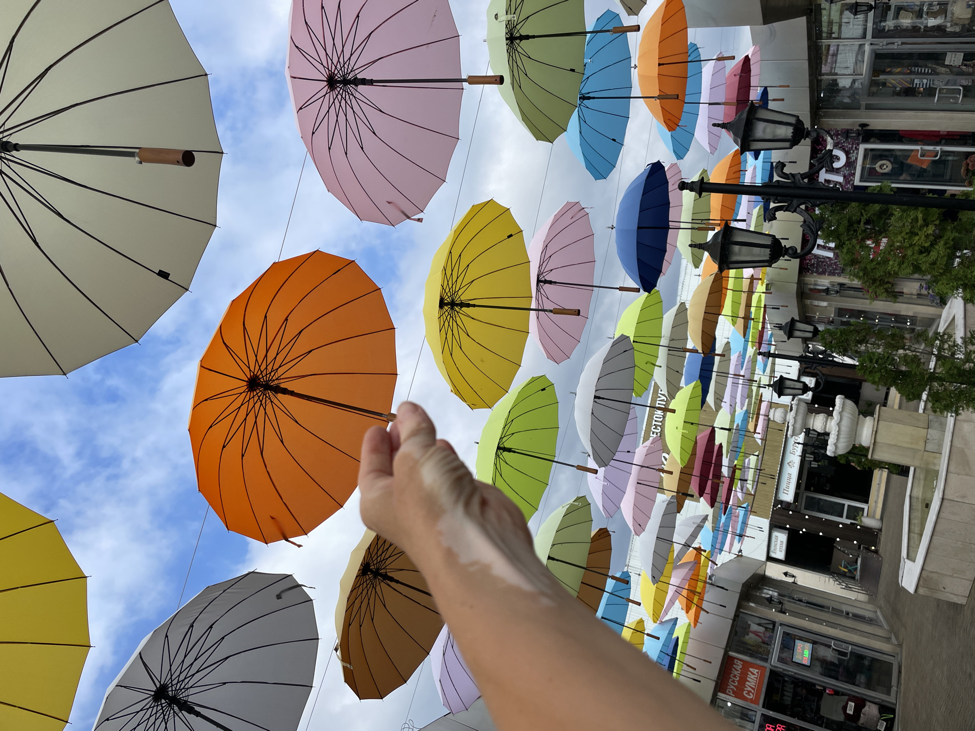 This media file features a vibrant outdoor scene, likely a street or pedestrian area, decorated with numerous colorful umbrellas suspended in the air. The umbrellas are in a variety of colors including pink, orange, yellow, green, blue, and white, creating a festive and eye-catching display. The view is from below, looking up towards the sky, which is partly cloudy. A person's arm and hand are prominently in the foreground, reaching up towards the umbrellas, suggesting an interaction with or appreciation of the installation.
The setting appears to be a commercial area with shops visible in the background, some with awnings and storefronts. Lampposts with ornate designs and a fountain are also present, adding to the ambiance of the location. Some text is visible on the storefronts. One sign reads "РУССКАЯ СУМКА" (Russian Bag) and another, more distant sign, has Cyrillic lettering that includes "ПИЦЦА" (Pizza) and "БУРГЕР" (Burger). The overall impression is one of a cheerful and decorated public space, possibly a tourist attraction or a place for a special event. The time of day seems to be daytime, with natural light illuminating the scene, and the weather appears to be overcast or partly cloudy. The emotion conveyed is one of joy and wonder, due to the colorful umbrellas and the engaging composition.