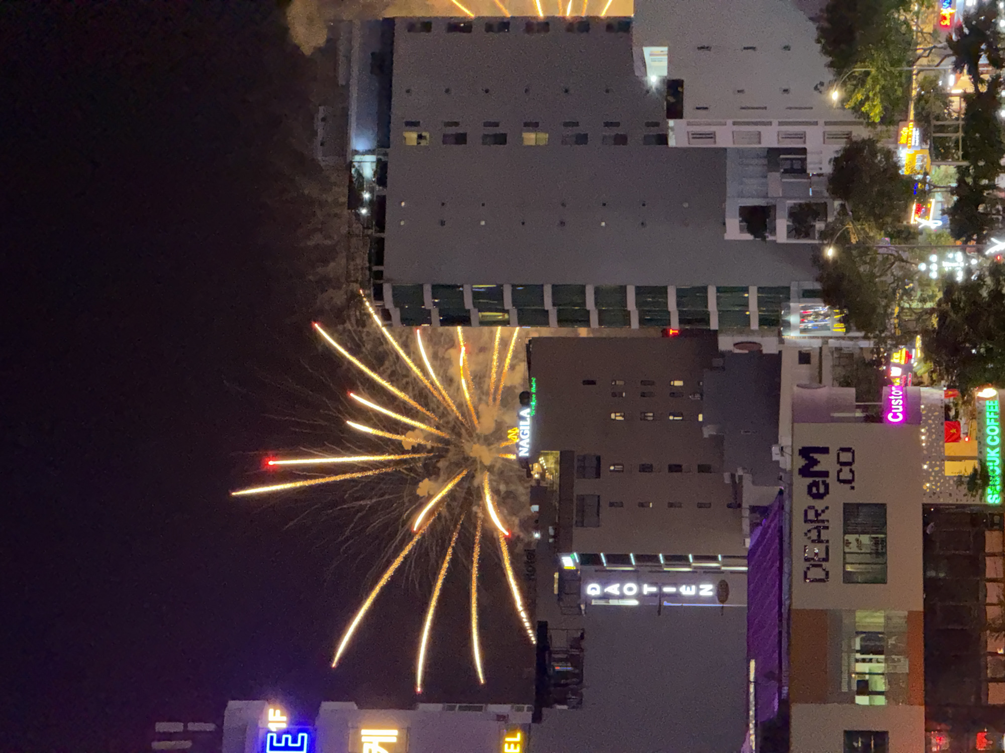 This is a nighttime photograph of a city skyline in Da Nang, Vietnam, featuring a fireworks display. The foreground is dominated by several buildings of varying heights. The closest building on the left has a purple roof and signage that reads "DEAR eM .CO". Below this, another building displays colorful lights and signage for "SEMGUK COFFEE". Further back, a taller, slender building has vertical neon signage that reads "DAO TIEN".
In the mid-ground, more buildings rise, including one with signage for "NAGILA". The fireworks are the most prominent element, with bright orange and yellow bursts exploding in the dark sky, casting light on the surrounding buildings. The overall scene depicts an urban environment during a celebration.