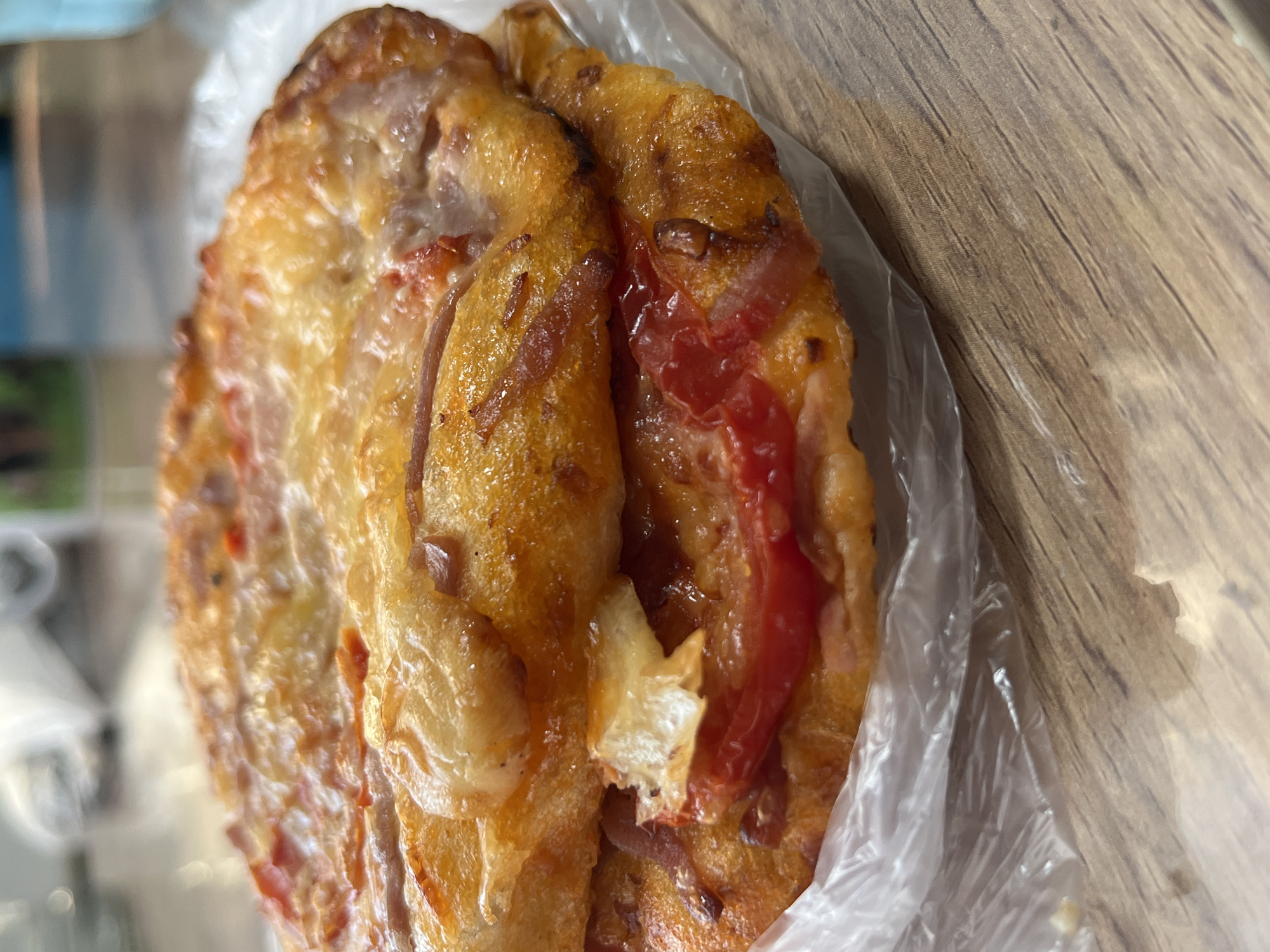 A close-up shows a round, golden-brown pastry item inside a clear plastic bag on a light brown wooden surface. The pastry appears to be a savory type, potentially a small pizza or pie, with visible toppings including cheese and what looks like red peppers or tomato. The setting may be in Nizhny Tagil, Russia, but there are no elements in the image that specifically confirm this location. The image is tightly framed on the food item and surrounding surfaces, with some blurred background elements that are unidentifiable.