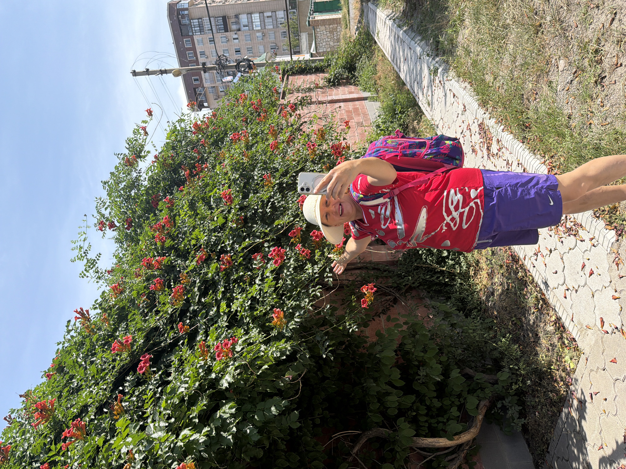 A smiling woman in a red t-shirt, white hat, and purple shorts takes a selfie with her phone in front of a wall covered in green leaves and red flowers. She has a pink and purple backpack on. To the right of the woman, a paved sidewalk runs along a grassy area. In the background, a tall, multi-story building and a utility pole with wires are visible. The sky is clear and blue.
