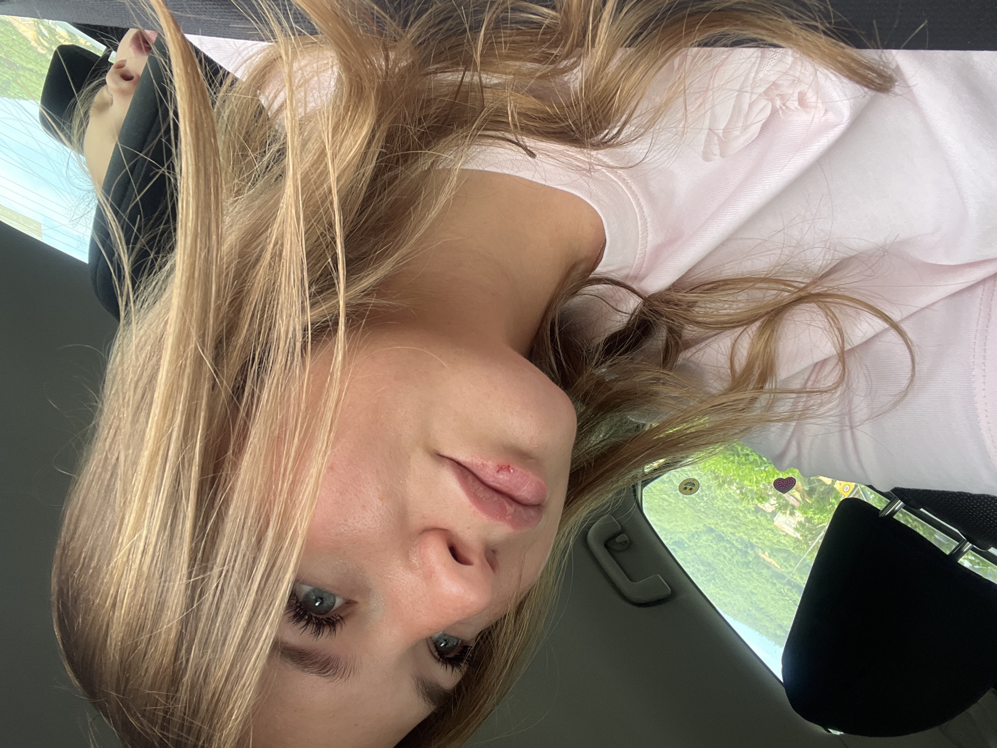 A young woman with blonde hair and blue eyes is inside a car. She is wearing a pink shirt and has a seatbelt on. There is a sticker on the car's window visible through the passenger side window showing green trees. The location cannot be determined from the image.