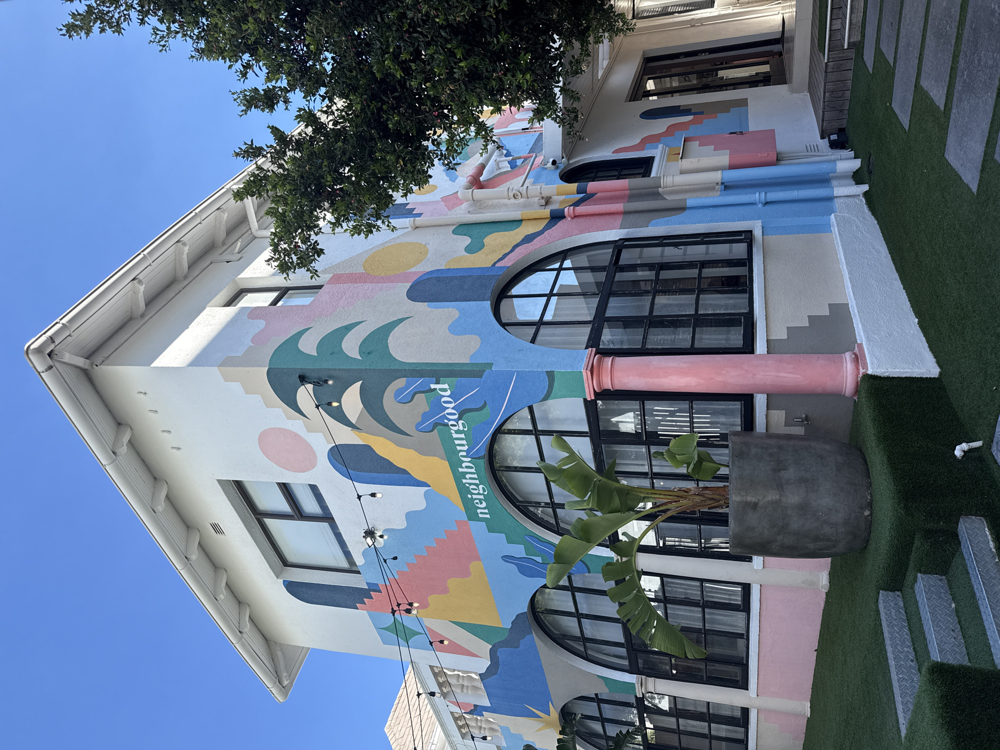 A low-angle outdoor shot depicts a three-story building in Cape Town, South Africa, distinguished by its vibrant mural. The building's facade is adorned with a colorful design that includes abstract shapes, waves, and a representation of the sun. The word "neighbourgood" is written within the mural. The building features arched windows with black frames. A large planter containing a tropical plant sits near the building's base. Strings of lights are hung on the side of the building. Above, a tree's foliage partially obscures the top of the building. The sky is visible in the upper left corner. A section of artificial turf is laid around the building's perimeter.