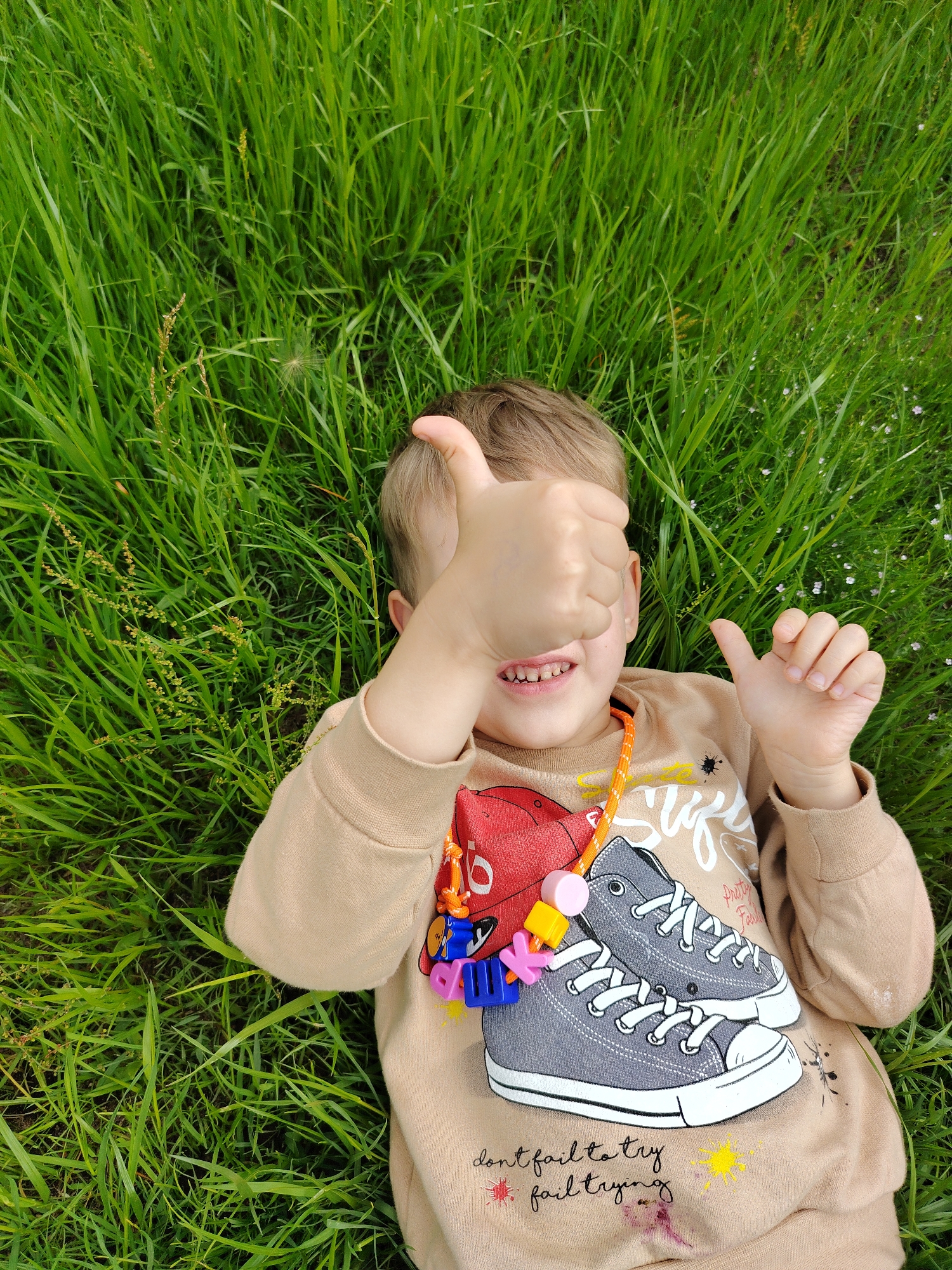 A young child lies on their back in a field of green grass, their head to the upper right of the frame. The child has light brown hair and is wearing a tan sweatshirt with a graphic design of two gray sneakers and text at the bottom. They are smiling and holding up two thumbs, with their right hand covering their eye. A colorful beaded necklace with charms, including what appears to be a red baseball cap, hangs around their neck. Small wildflowers are scattered throughout the grass. The image appears to have been taken outdoors during the day, with natural light. The child's expression and thumbs-up gesture suggest a feeling of happiness or satisfaction. The text on the sweatshirt reads: "don't fail to try fail trying."