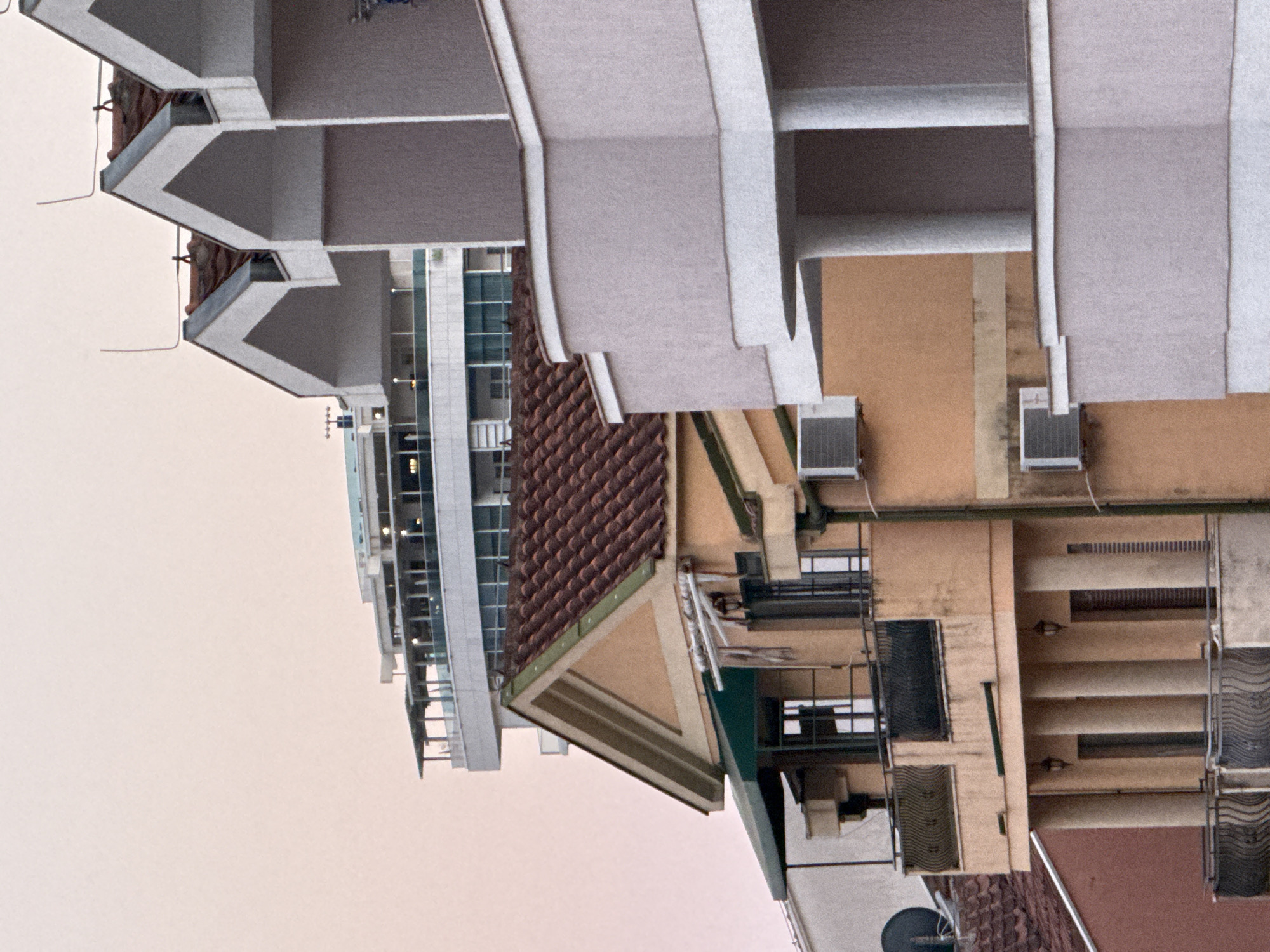 The image captures a cityscape in Budva, Montenegro, focusing on the facades of several apartment buildings. The sky is a pale, muted pink, suggesting either dawn or dusk. In the foreground, a light orange building with a red tiled roof features balconies with ornate metal railings and is adorned with air conditioning units. To the right, a lighter colored building with curved balconies and prominent white trim is visible, with sharp, angular white roof elements extending upwards. Behind these structures, a modern, multi-story building with large windows and a balcony deck stands out. The overall scene depicts a dense urban environment with a mix of architectural styles. There are no people visible in the image.