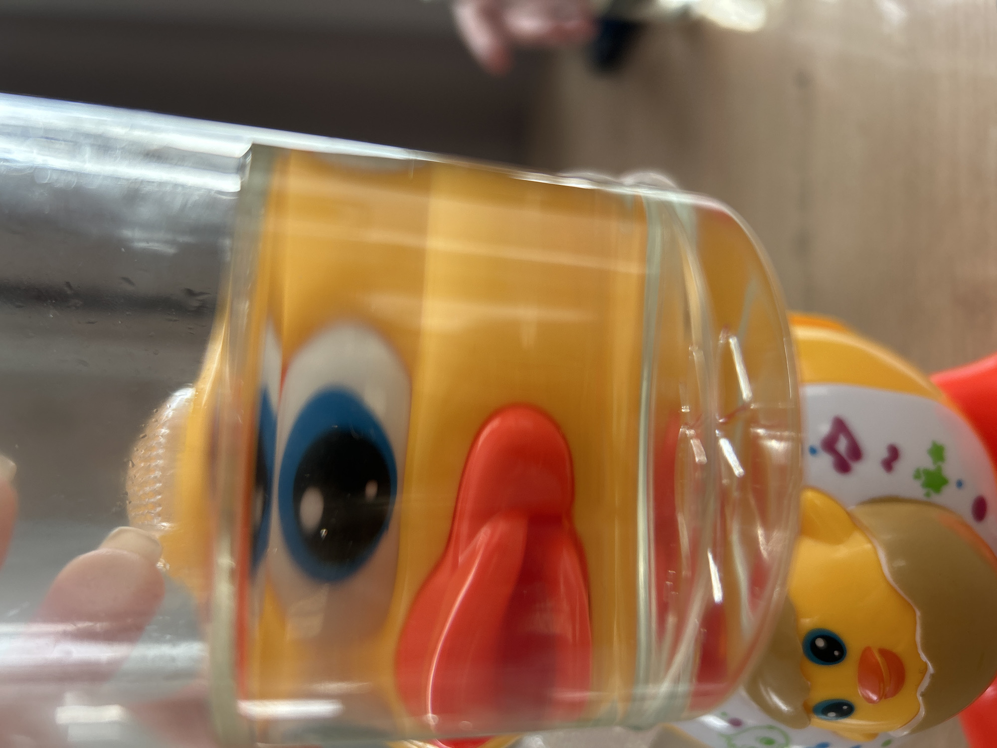 The image displays two toy characters, primarily visible within a clear, cylindrical container. The photograph is rotated clockwise, presenting the container horizontally.
The main subject is a vibrant yellow toy, possibly a duck or similar animal, featuring a large blue eye with a black pupil and a prominent red beak or mouth. This toy is partially obscured by the clear container, which shows reflections and slight distortions of its surface. Small water droplets or condensation appear on the inner surface of the container near this toy.
To the right of the main toy, another smaller yellow toy character is visible, partly inside and partly outside the clear container. This second character has a brown, possibly molded, "hair" or hat, small blue eyes with black pupils, and an orange beak. Its body is white and adorned with colorful patterns including purple musical notes, green abstract shapes, and blue dots.
The container itself is transparent, allowing the toys inside to be seen clearly while also reflecting ambient light. The background is blurred and indistinct, suggesting an indoor setting with varying shades of brown and beige. A very faint, blurred pinkish shape in the foreground or reflected on the container might be a human hand or fingers, potentially interacting with or holding the container. No specific activity is depicted beyond the static presence of the toys within the container. There is no visible text in the image. The time of day and weather are not discernible.