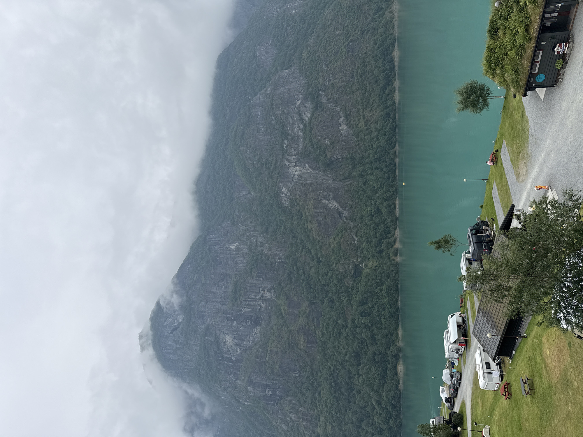 The image depicts a campsite situated by a tranquil, turquoise-colored lake, with a formidable, cloud-shrouded mountain range serving as a dramatic backdrop. The campsite is equipped with various recreational vehicles, including caravans and motorhomes, parked on a gravel and grassy terrain. Several picnic tables are scattered throughout the area, suggesting a recreational setting. A small, dark-colored building with a grass-covered roof is visible on the right side of the frame, adding a rustic charm to the scene. Two individuals are present, one seated at a picnic table near the lake and another standing near a building. The overcast sky contributes to a serene and somewhat moody atmosphere. The location is identified as Olden, Norway, a village nestled within a picturesque fjord landscape, renowned for its natural beauty and outdoor activities.
