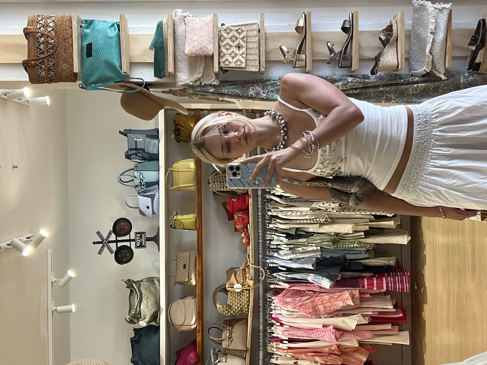 A young Caucasian woman with blonde hair takes a selfie in a boutique clothing store. She is wearing a white tank top with embellishments and a white skirt. She is also wearing several bracelets and a necklace. She is holding a smartphone in her right hand. Behind her, shelves are stocked with handbags, hats, and shoes, and clothing racks are filled with garments. The store appears to be in Pollença, Spain, given the distinctive architectural style of the building and the overall ambiance.