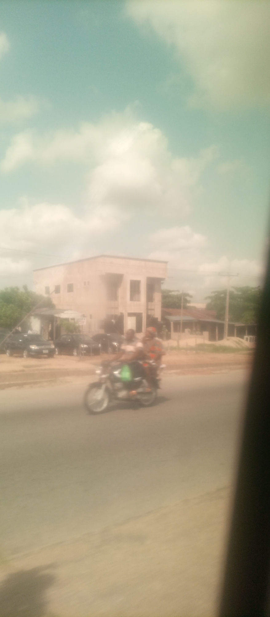 The image depicts a street scene in Jalingo, Nigeria, characterized by a building and a moving motorcycle. The sky is a pale blue with scattered white clouds, suggesting daytime. In the background, a two-story building with a light-colored facade and dark windows stands prominently. To the left of the building, green foliage is visible. In the mid-ground, a motorcycle is in motion on a paved road. Two individuals are seated on the motorcycle; the driver appears to be wearing a light-colored shirt and the passenger, a darker shirt with orange accents, and is wearing a helmet. A green bag is visible strapped to the motorcycle. Several cars are parked to the left of the motorcycle, near the building. The overall image has a slightly blurred quality, possibly due to motion or the camera's focus.