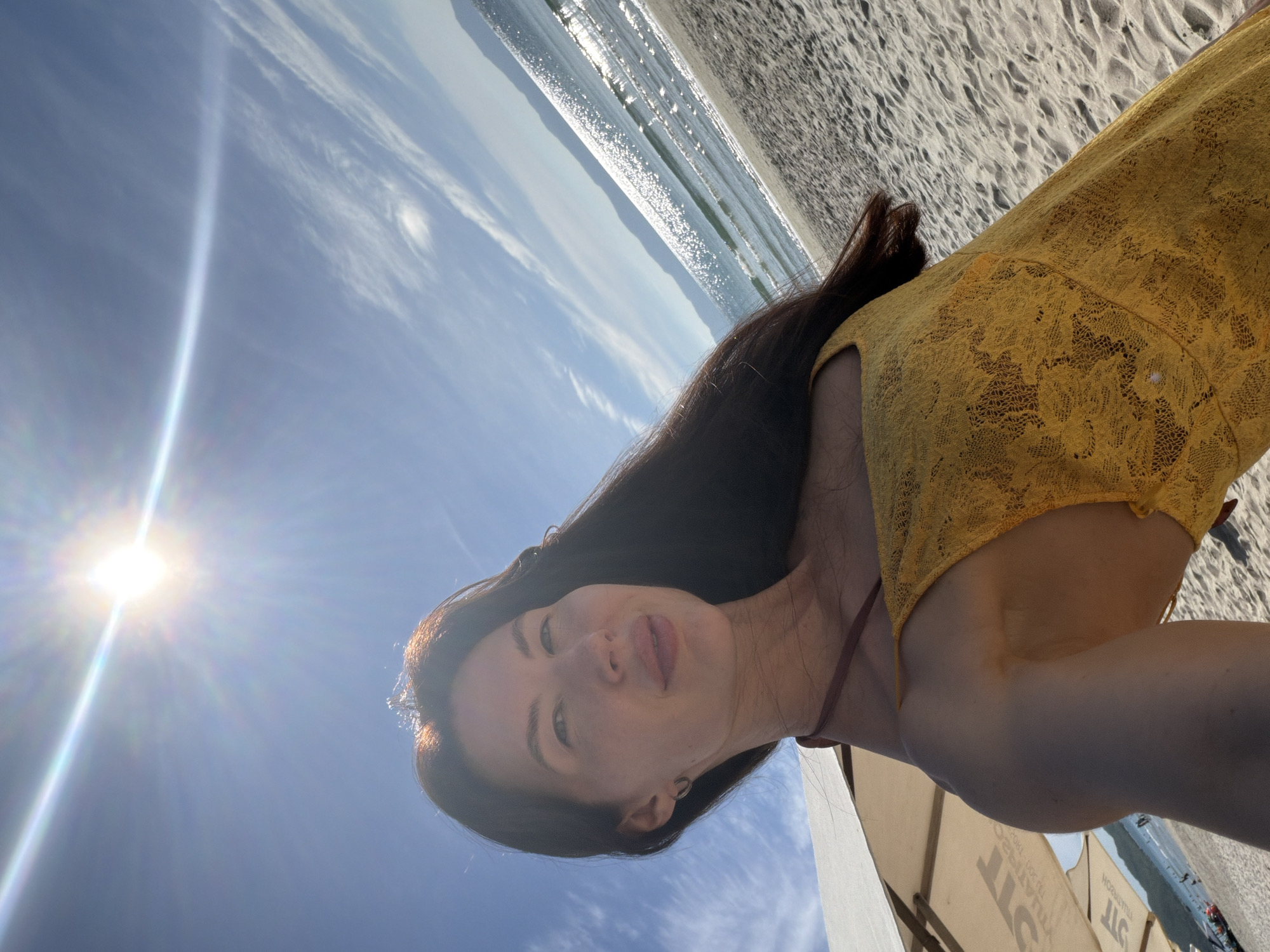 A woman is taking a selfie on a sandy beach. The sun is bright and positioned in the upper left corner of the image, creating lens flare. The woman has long, dark hair and is wearing a yellow lace dress. She is looking towards the camera with a slight smile. In the background, the ocean with gentle waves is visible, and a coastline with hills can be seen in the distance. The sky is blue with some wispy clouds. Beach umbrellas are present on the left side of the frame. The scene appears to be a sunny day at the beach.