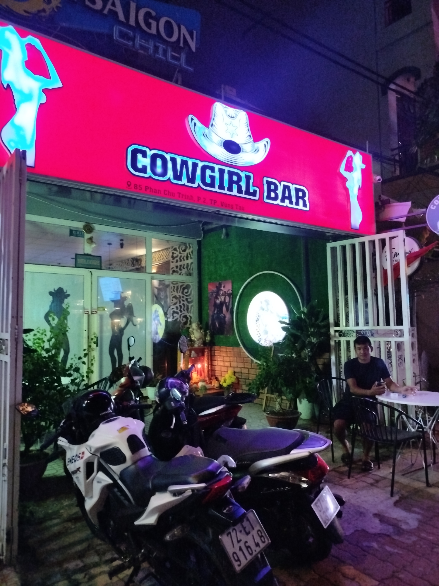 The image shows the exterior of a bar named "Cowgirl Bar" located at 85 Phan Chu Trinh, P.2, TP. Vung Tau. The sign is bright red with the name in blue letters, and the silhouettes of women are featured on either side. A man is sitting at a table outside the bar, and several motorcycles are parked in front of the entrance. The environment suggests it is nighttime.