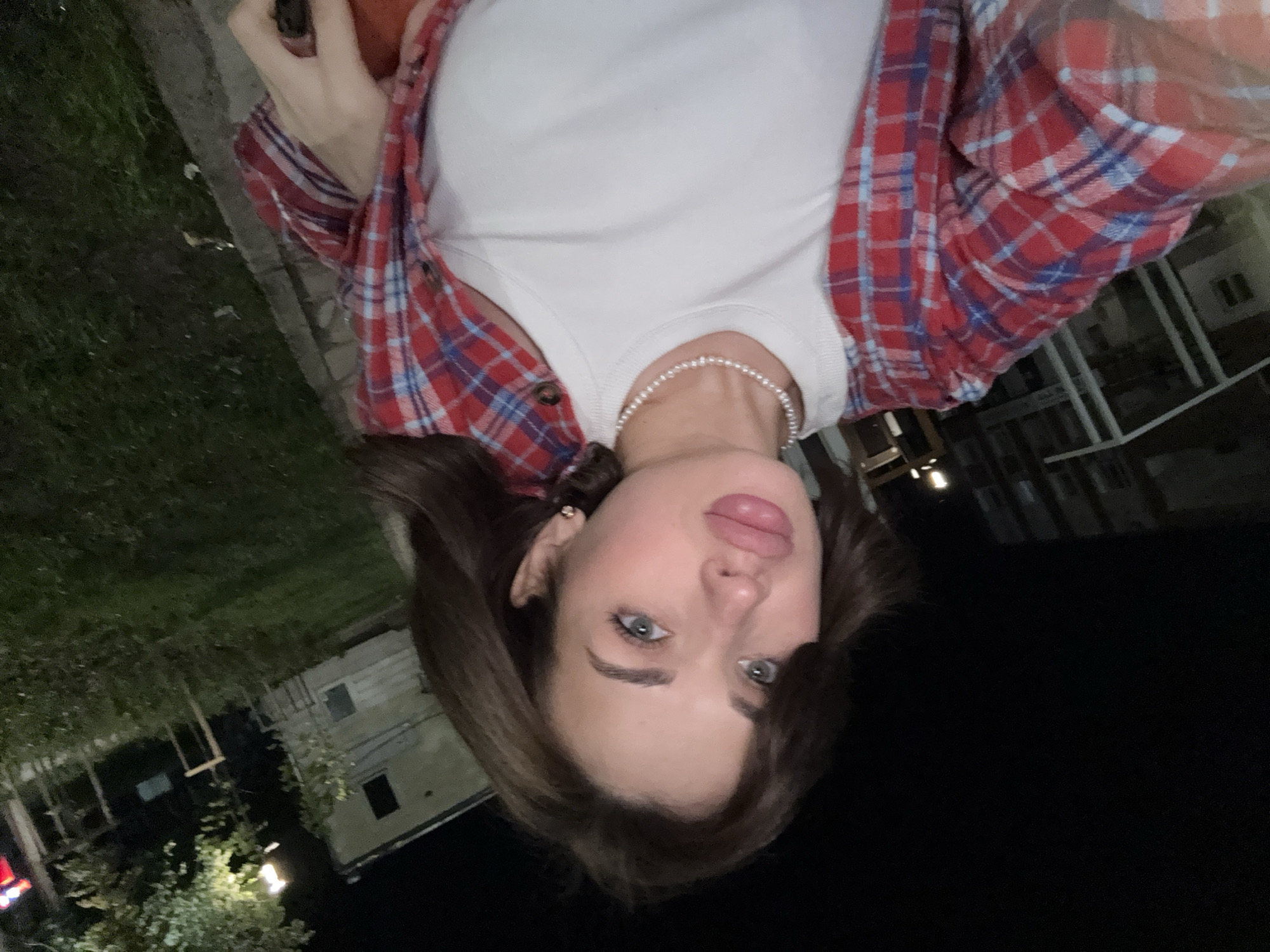 A woman with brown hair and blue eyes is taking a selfie outdoors at night. She is wearing a white T-shirt, a pearl necklace, and a red and blue plaid flannel shirt. The background shows buildings and trees, indicating an urban setting. The photograph was taken in Sarapul, Russia.