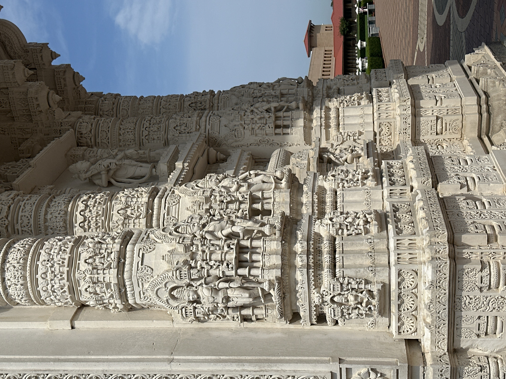 This is a close-up, eye-level, outdoor, daytime, medium shot of the intricately carved white marble facade of the BAPS Shri Swaminarayan Mandir in Bartlett, Illinois, United States of America. The image focuses on the detailed relief carvings on a massive stone pillar, depicting various figures and decorative motifs. The pillar has multiple tiers, each adorned with smaller sculptures of deities and intricate patterns. The scale of the carvings suggests religious or cultural significance. In the background, a modern building with a terracotta roof is visible, along with a paved walkway and manicured green spaces. The sky is a clear blue with a few wispy clouds. The lighting is bright, indicating a sunny day, casting subtle shadows that highlight the depth and texture of the carvings. No people are clearly visible in the frame. The overall impression is one of architectural grandeur and meticulous craftsmanship.