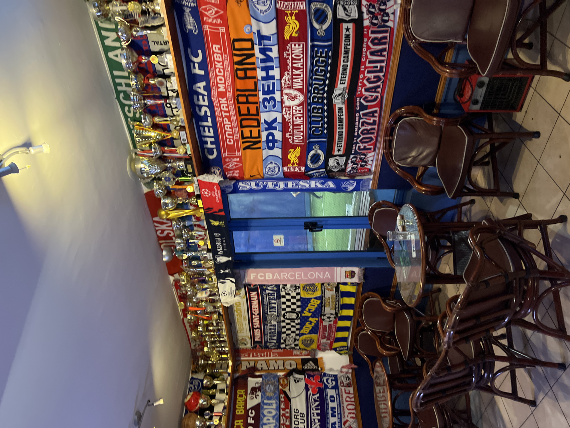 The image depicts the interior of a bar in Budva, Montenegro, identifiable by the presence of a "Forza Cagliari" scarf and other European football team scarves displayed along the walls and above a doorway. These include Chelsea FC, Spartak Moscow, and FC Barcelona. Additionally, numerous trophies are lined up on shelves next to the scarves. A stack of dark wood chairs is placed near tables with glass tops. The scene suggests the bar's interior features football memorabilia and seating for patrons. Through a window, it is possible to see the outside of a football pitch. An electric heater is also visible on the floor next to the seating.
