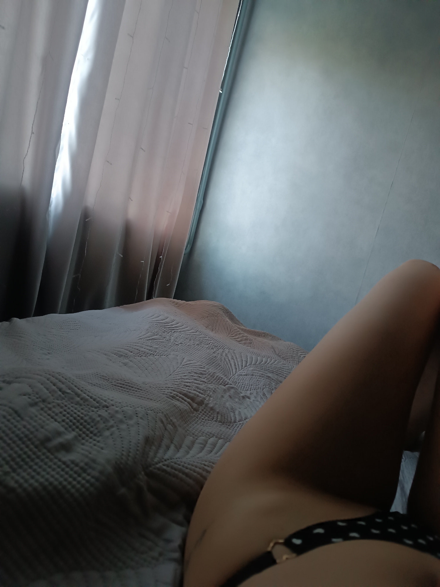 A high-angle indoor shot shows a person reclining on a bed in a room. A white comforter with a quilted pattern covers the bed. The lower half of a person is visible in the right foreground, wearing black underwear with white polka dots. Behind the bed and person is a wall and a window covered by a white curtain. The location is unknown.