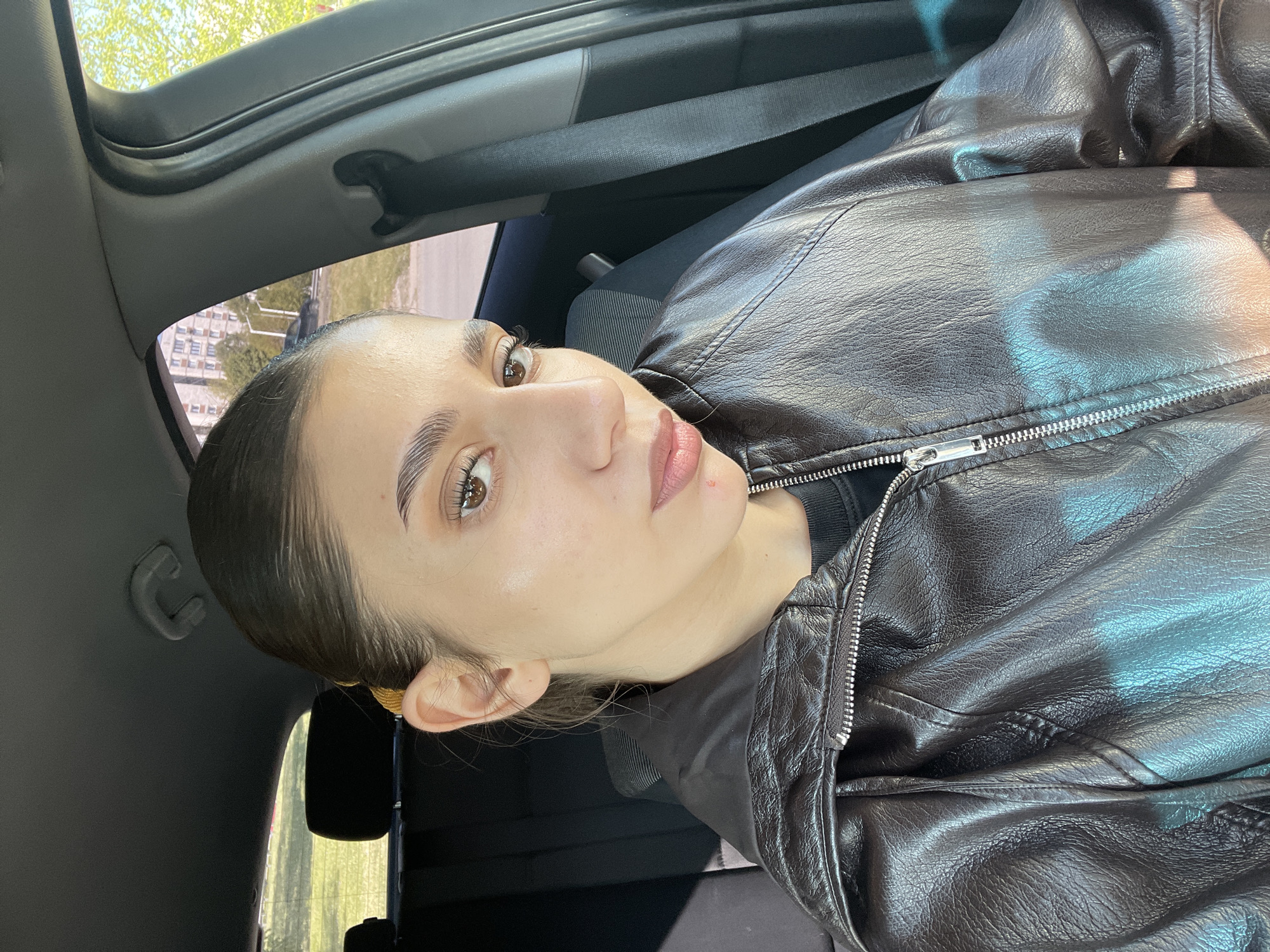 A light-skinned person with brown eyes and dark hair pulled back is in the driver's seat of a car. They are wearing a dark brown leather jacket. The interior of the car is visible, including the ceiling, seatbelt, and headrest. Outside the car window, there are blurred trees and buildings, suggesting an urban environment. It is not possible to determine the exact location in Sofia, Bulgaria, based solely on this image.