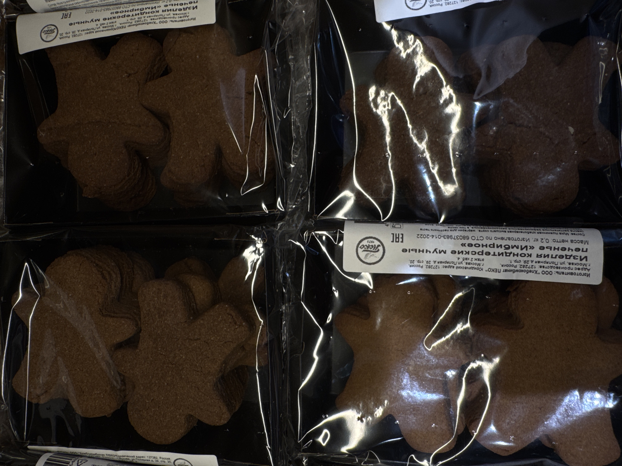 Four packages of gingerbread cookies are displayed in a grid formation. Each package contains multiple cookies in the shape of a gingerbread man. The cookies are a dark brown color and appear to be individually wrapped or layered within the transparent plastic packaging. Labels are visible on the right side of the packages. One label, in Russian, states "Изготовитель: ООО "Хлебокомбинат ПЕКО" Юридический адрес: 127282, Россия, г.Москва, ул.Полярная д.29, стр. 20, этаж 2 каб. 4." This translates to "Manufacturer: OOO "Khlebokombinat PEKO" Legal address: 127282, Russia, Moscow, Polyarnaya St. 29, bldg. 20, floor 2, office 4." Another line on the label reads "Изделия кондитерские мучные печенье «Имбирное»," meaning "Flour confectionery products gingerbread cookies." The image is a close-up shot, suggesting it might be taken in a retail environment, such as a grocery store or bakery. The lighting creates reflections on the plastic packaging.
