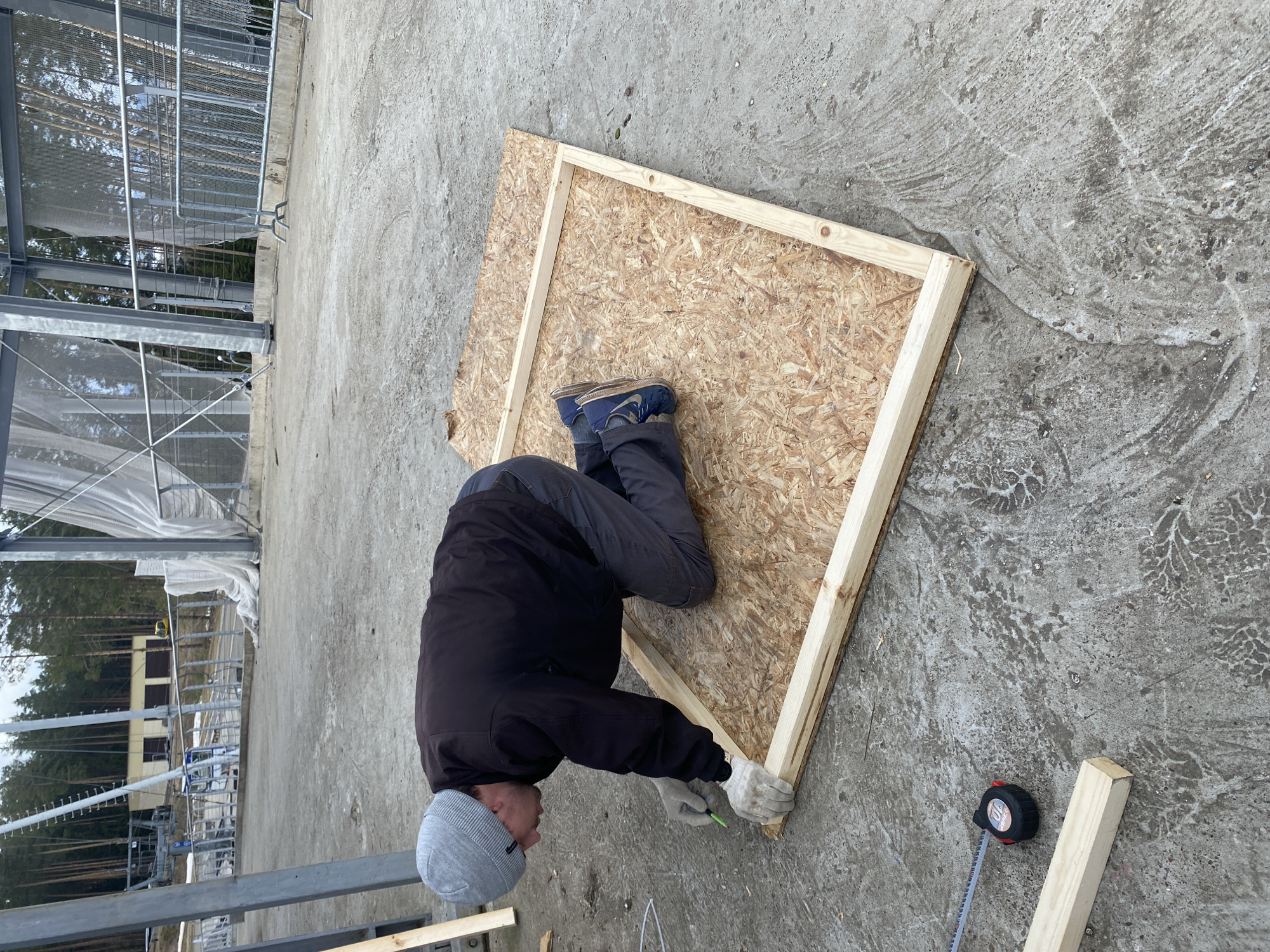 An outdoor, eye-level shot shows a person actively engaged in construction or woodworking on a concrete ground. The individual, dressed in a gray beanie, a dark jacket, dark trousers, and light-colored work gloves, is crouched over an Oriented Strand Board (OSB) panel. This OSB panel has a wooden frame partially constructed around its edges. The person is using a green tool, likely a marker or pencil, to work on one of the wooden frame pieces. Their blue and gray sneakers are visible on the OSB board.
A black and yellow Stanley tape measure is lying on the concrete to the right of the person, with its tape extended, showing "10m" and "STANLEY" printed on it. Loose wooden planks are also scattered on the concrete surface. The ground itself is a rough, textured concrete, with some visible patterns.
In the background, to the left, metal structures, possibly part of a large construction or industrial framework, are prominent. Beyond these structures, a dense line of tall evergreen trees is visible under a bright, possibly overcast, sky. A light-colored building can be seen nestled among the trees in the distance. The scene suggests a construction site or a workshop area in an outdoor setting in Прикамский, Russia, during daytime.