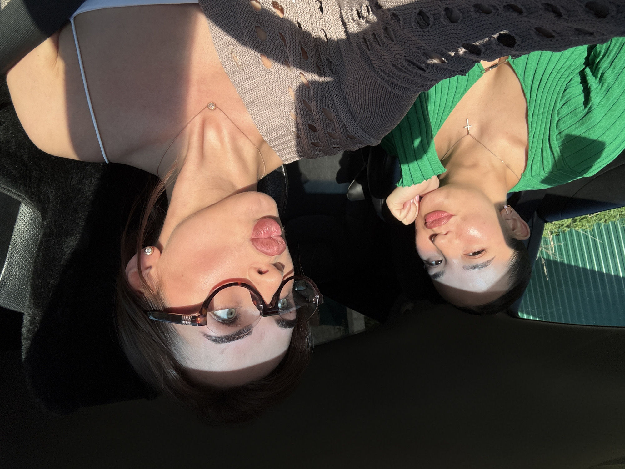 Two women are in a car. One is wearing glasses and a tan sweater, the other is wearing a green shirt and a silver cross necklace. The location is Perm, Russia. A fence can be seen in the background through the window.