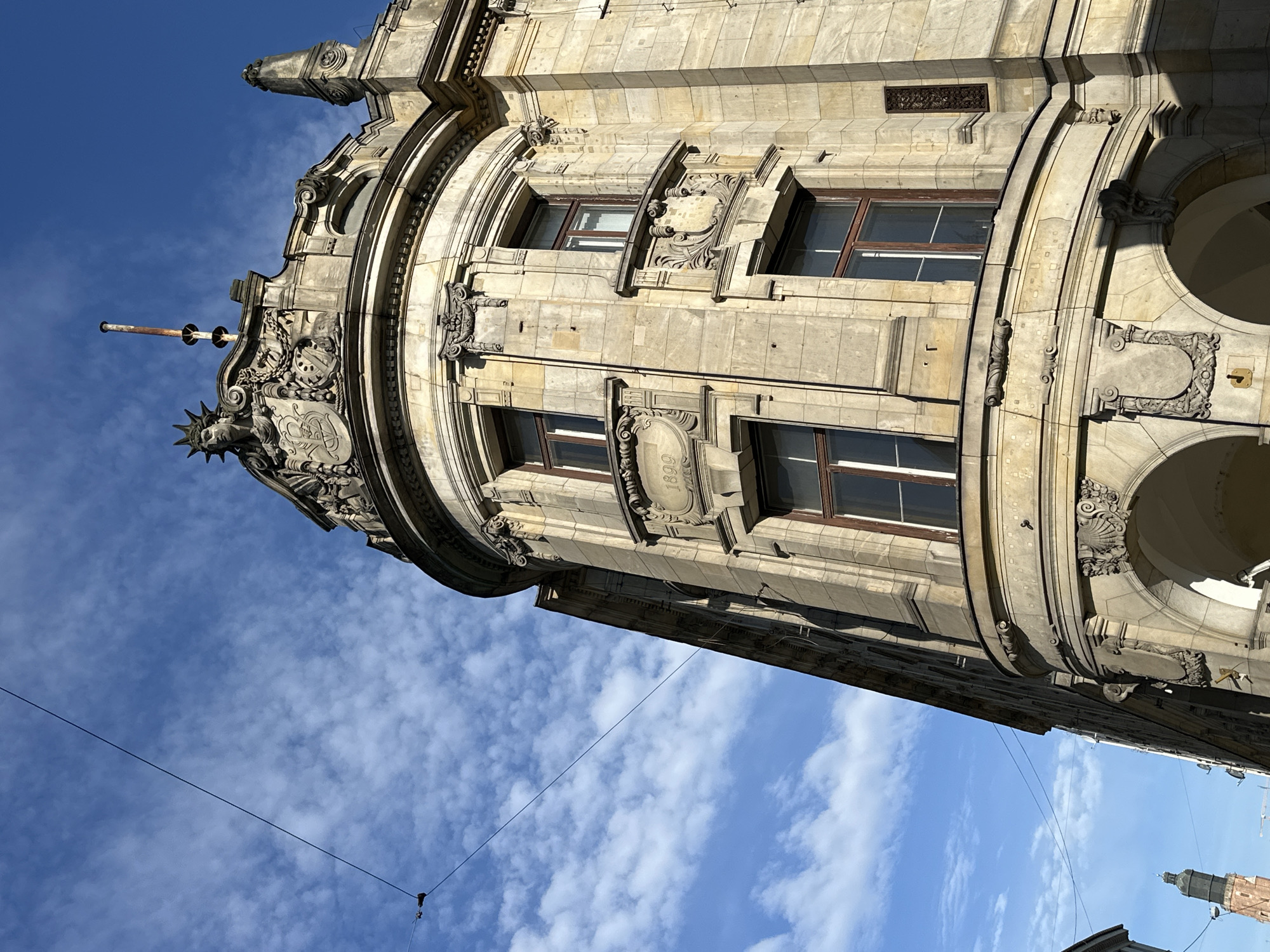 The image shows a close-up, low-angle view of a historic building against a partly cloudy, blue sky. The building features ornate architectural details, including arched windows, decorative moldings, and carvings. One prominent feature is a plaque inscribed with the number "1898."
The building is located in Wrocław, Poland.