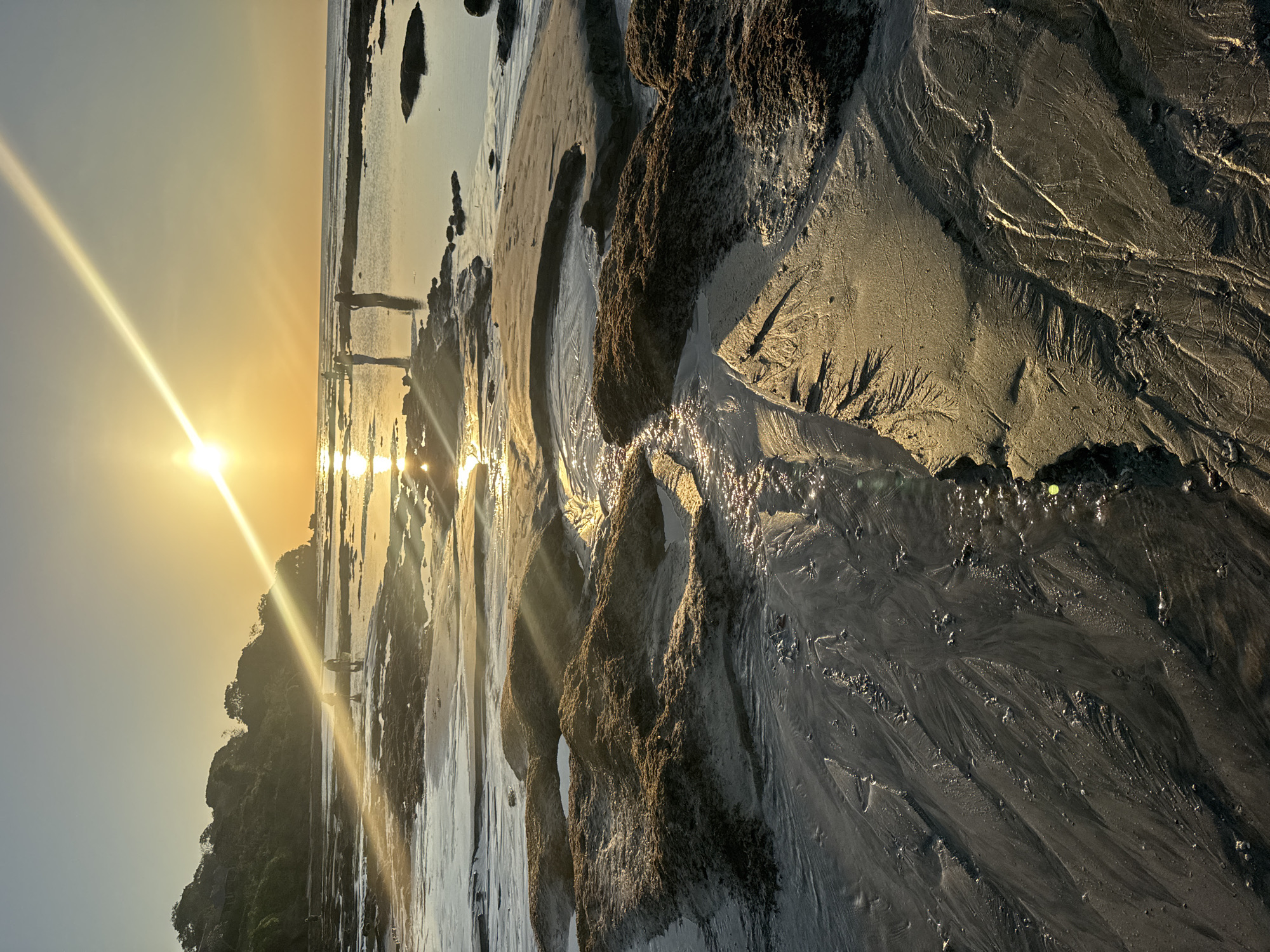 The image depicts a coastal scene at sunset in Kuta Selatan, Indonesia. The sun, low on the horizon, casts a bright glare and long rays across the wet sand and shallow pools of water. Rocky outcrops, covered in dark seaweed, punctuate the sandy shoreline. Several people, appearing as silhouettes against the bright sky, are scattered across the beach and in the water. The terrain is uneven, with rippling sand patterns formed by receding water. Vegetation and a small building are visible on a gentle slope in the background. The overall atmosphere is serene, illuminated by the warm glow of the setting sun.