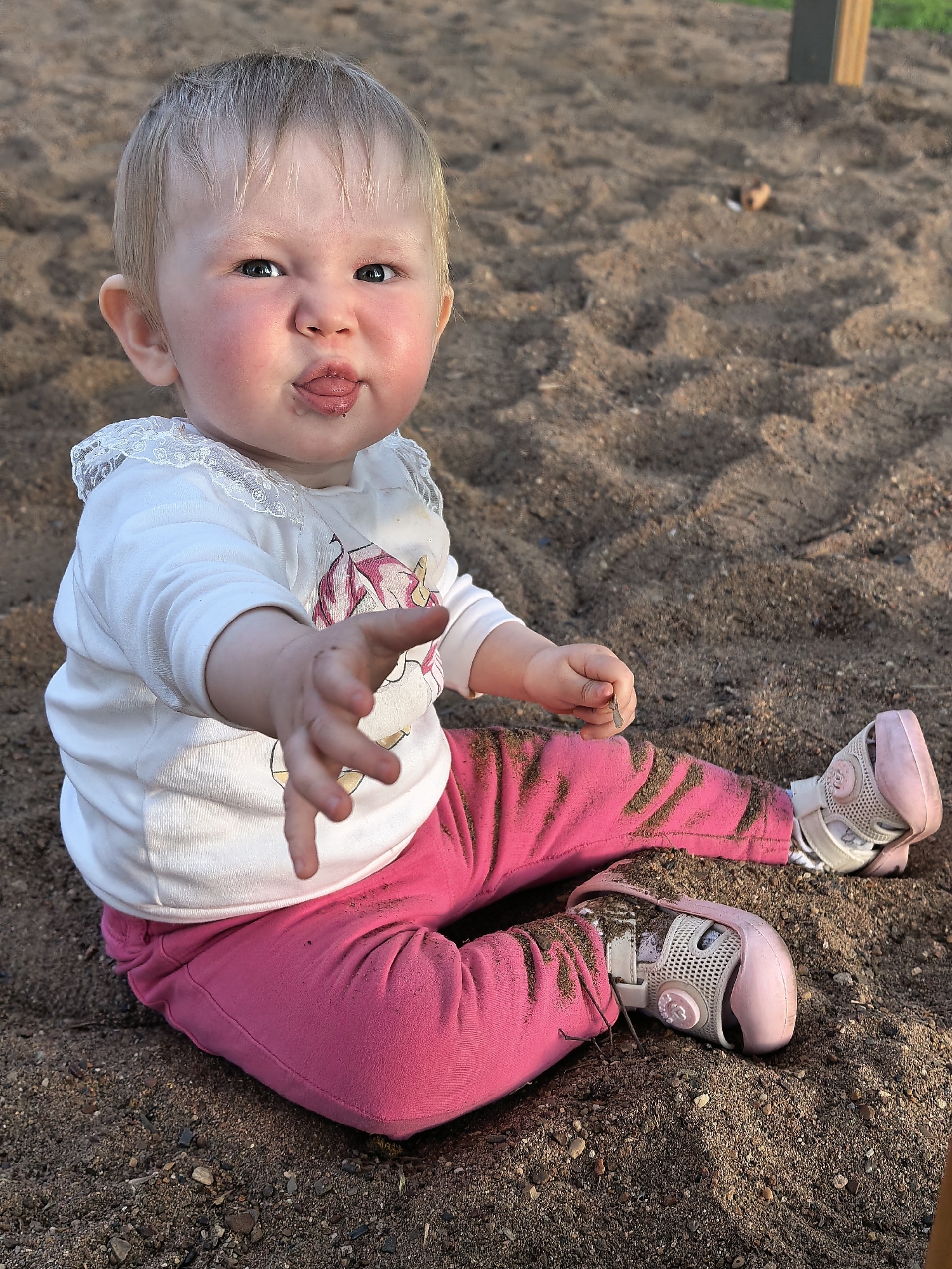 A baby sits in a sandy area with some small rocks. The baby has pink pants, pink and white shoes, and a white, long-sleeved shirt with lace at the neck and a unicorn design on the front. The baby is sticking out its tongue and holding out its right hand. The pants and shoes are covered in sand. The background consists of sand and part of a wooden post. The location is Sterlitamak, Russia.