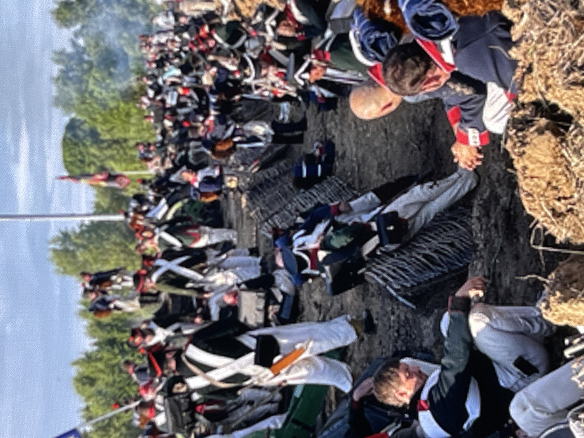 The image shows a historical reenactment, likely of a Napoleonic-era battle, with participants dressed in period military uniforms. The scene depicts a group of individuals in a rough, outdoor setting with a dirt ground and scattered debris. Many are wearing white pants, dark jackets with decorative elements, and tall hats. Some are standing, while others are seated or lying down. There is a sense of chaos or aftermath, with some figures appearing to be injured or resting. Flags and barricades are also visible, suggesting a staged combat scenario. The background consists of trees and a partly cloudy sky, indicating an outdoor event. The image does not provide any specific information about the city of Moscow, Russia.