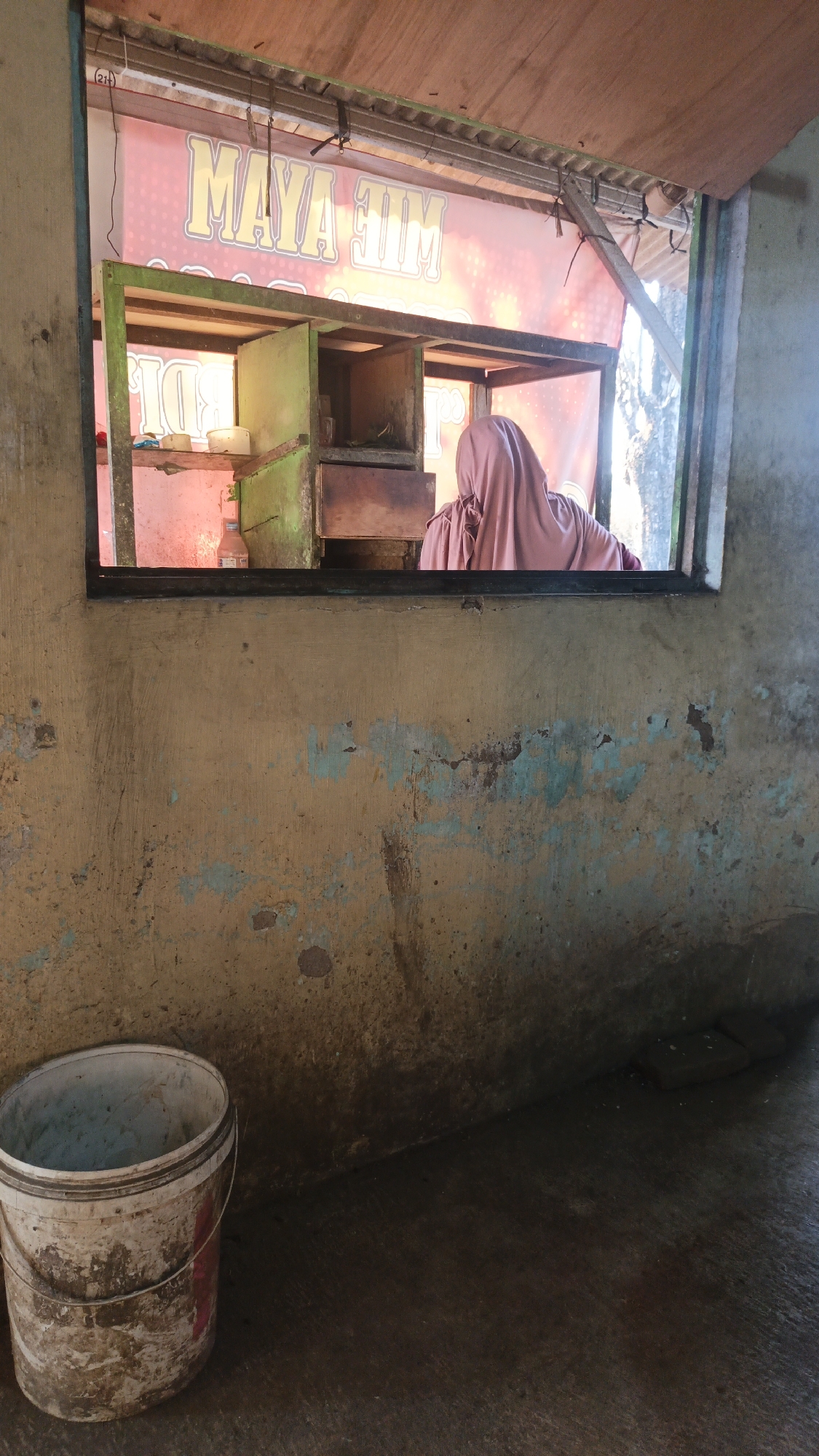 A person wearing a pink headscarf is seen from behind, standing at a food stall window. The stall has a sign with yellow lettering that reads "MAYA JIM MIE AYAM." Inside the stall, there are shelves and what appears to be cooking equipment. In the foreground, to the left, is a dirty white bucket. The wall of the stall is weathered and stained, with chipped paint in various colors. The floor is concrete. The scene appears to be outdoors, possibly in a rural or semi-urban setting within the city of Dawe, Indonesia, given the food stall's signage.