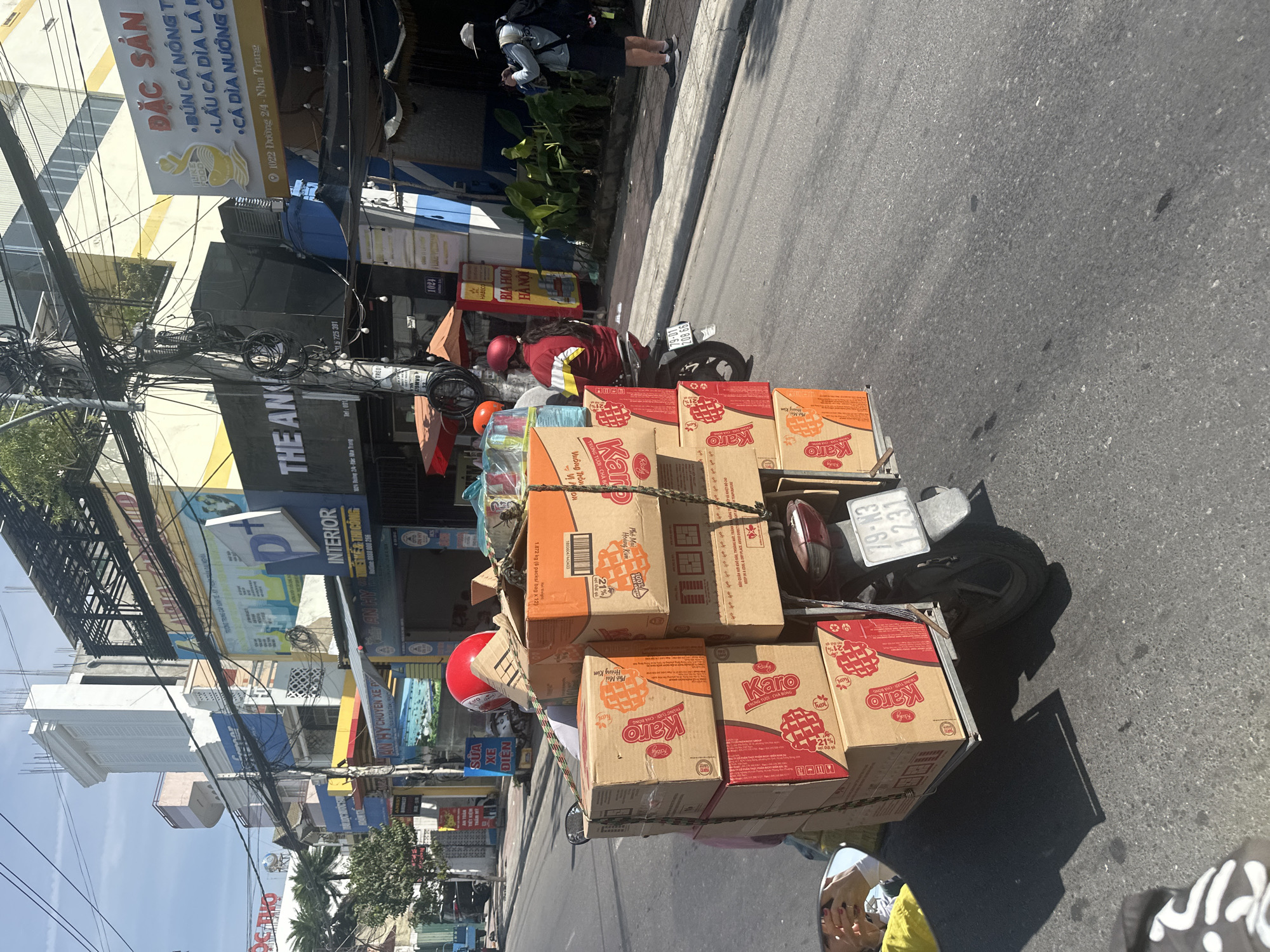 In Bac Nha Trang Ward, Vietnam, a motorcycle is laden with stacked cardboard boxes of "Karo" waffles and other goods, secured with rope. A person wearing a red helmet and a red and yellow jacket rides the motorcycle. The scene takes place on a city street with buildings lining the right side, displaying various signs including "THE AN", "INTERIOR", and a sign in Vietnamese advertising "ĐẶC SẢN" (Specialties). The sky is blue with some clouds. The street is paved and appears to be in a commercial area with overhead electrical wires present.