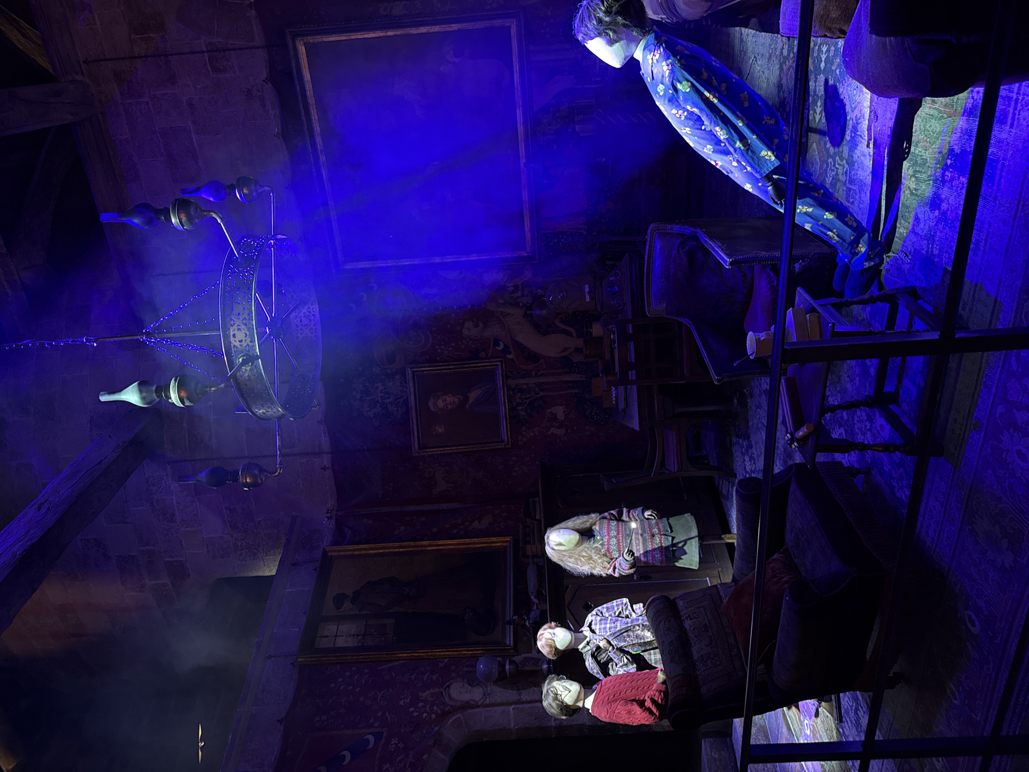 The image depicts a dimly lit room, possibly a recreation of a scene from the Harry Potter universe, with a strong blue lighting effect and atmospheric smoke. In the foreground, a dark armchair with a red cushion is visible. To the right, a figure in blue pajamas with a floral pattern is positioned at an unnatural, levitating angle, supported by a stand. This figure appears to be a mannequin.
In the mid-ground, three child-like mannequins stand together. The mannequin on the left wears a red sweater, the middle one a plaid shirt, and the right one a striped sweater and skirt. The mannequin on the far right holds a wand. Behind them, a dark wooden cabinet is laden with books and other objects.
The room is decorated with ornate tapestries on the walls and several framed portraits. A large, elaborate chandelier hangs from the ceiling. Furniture such as chairs and tables are present, suggesting a living or common room setting. The overall atmosphere is theatrical and immersive, likely a display in a museum or attraction. The image is taken from a slightly elevated perspective, looking down into the scene. The location of this scene is stated to be within the city of Three Rivers, United Kingdom.