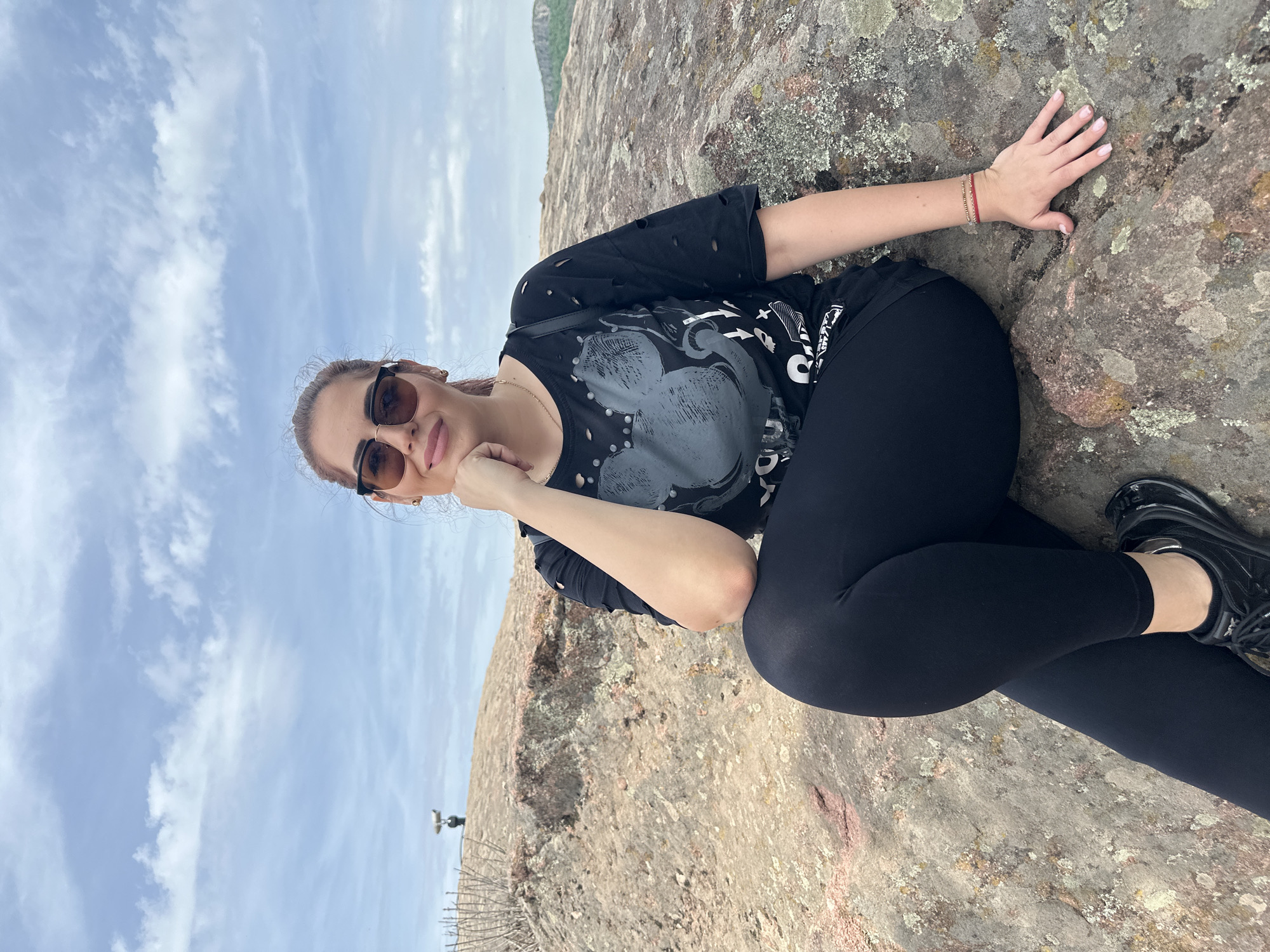 A woman is lying on a rocky surface in Belogradchik, Bulgaria. She is wearing sunglasses, a black t-shirt with a graphic, and black leggings. The sky is visible in the background with patchy clouds. In the distance, there is a mountain range. A metal fence and post are in the lower left corner of the image.