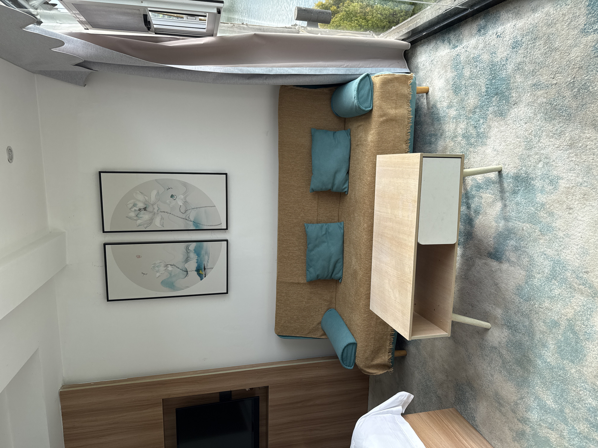 This interior scene shows a bedroom or sitting room with a combination sofa bed, side table, and artwork. The sofa bed has a brown woven texture with blue cushions and is positioned beneath a large window. A wooden side table with a white upper section sits to the side of the sofa. On the wall, two framed pieces of art featuring a floral or botanical motif hang above a wooden console table which has a black television. The location is potentially in Zhuoshui, China, but without additional details or context, this cannot be verified.