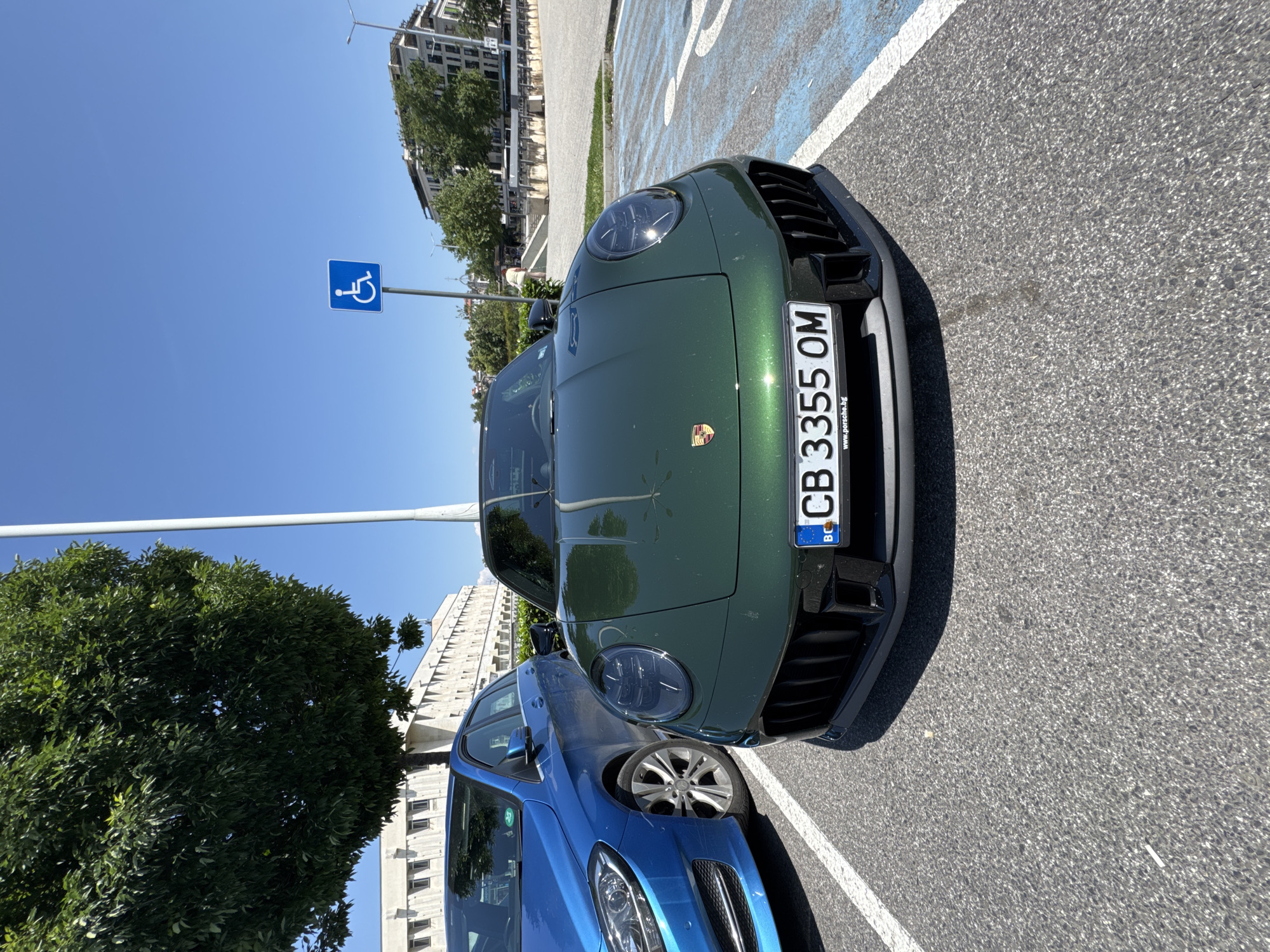 A green Porsche sports car is parked in a designated handicap parking space, as indicated by the handicap symbol on a signpost next to the car, in Plovdiv, Bulgaria. The car's license plate reads "CB 3355 OM". A blue car is parked adjacent to the Porsche, and urban buildings and trees are visible in the background under a clear blue sky.