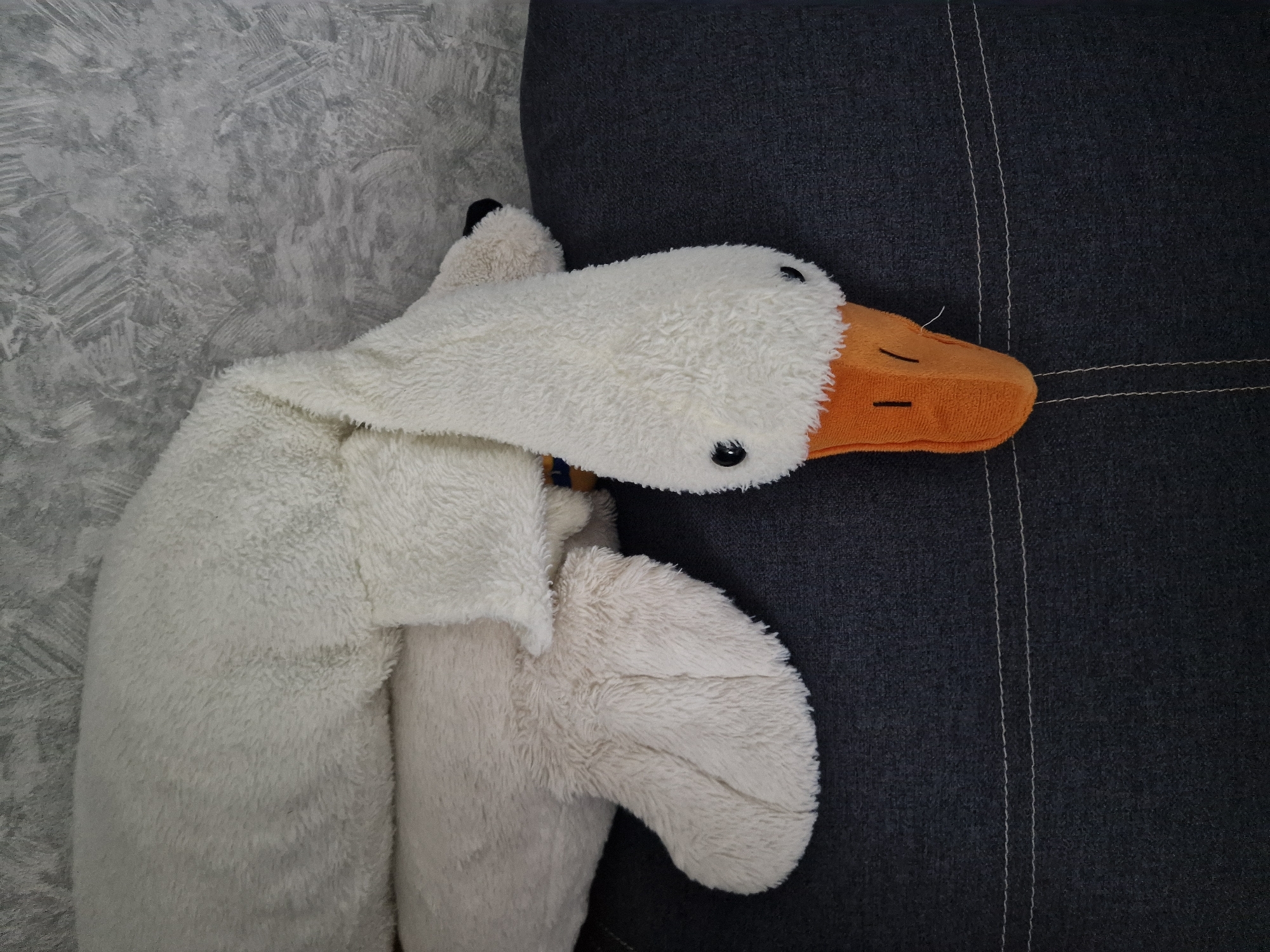 The image shows a plush white goose toy with an orange beak and black eyes. It is lying on a dark gray upholstered surface, possibly a couch or chair, with a textured gray wall in the background. The goose's body is elongated, and its head is bent downwards, with the beak resting on the surface. The lighting is neutral, and there are no people or other animals present. The location of Kursavka, Russia is not visually identifiable in the image.