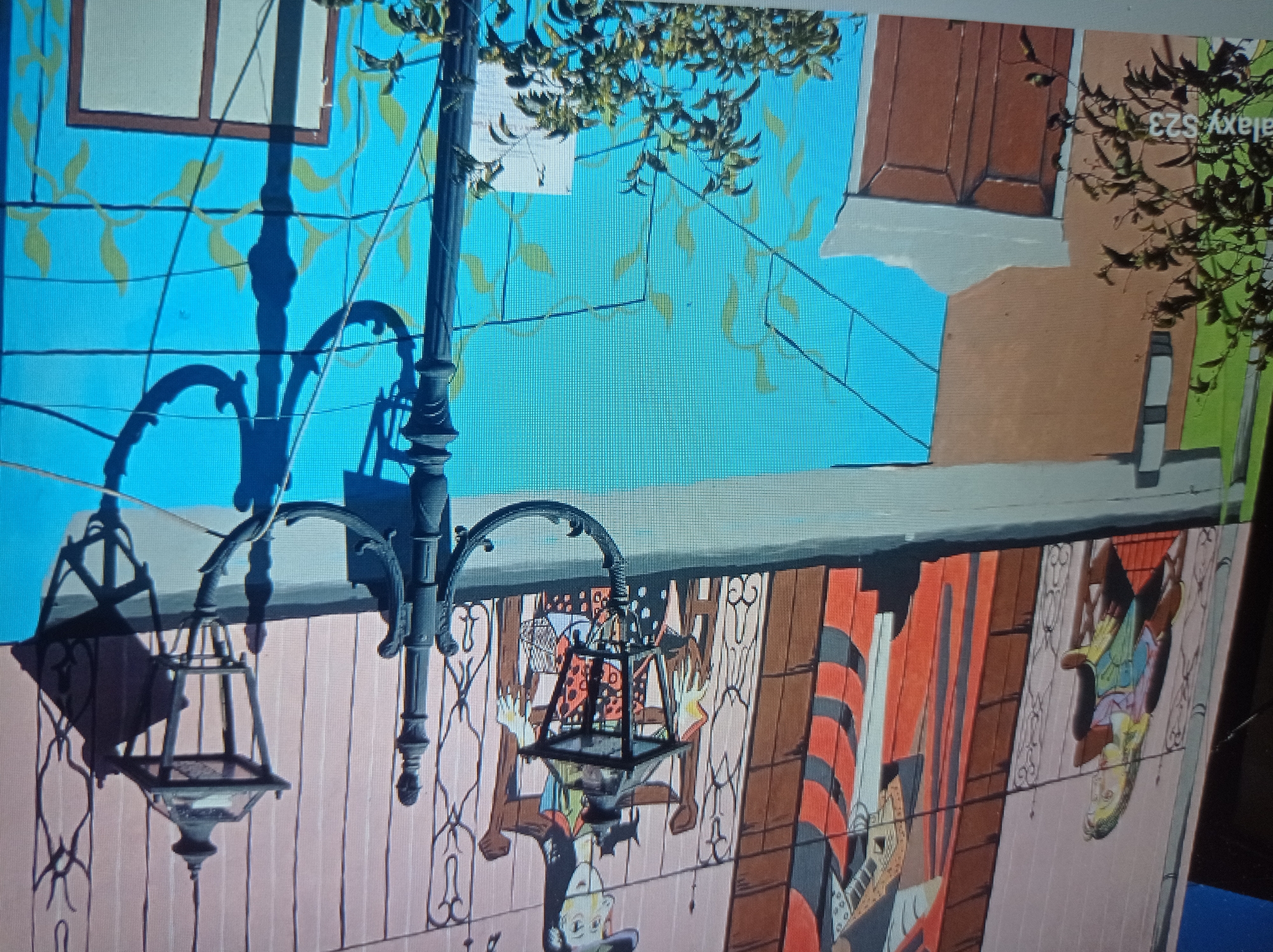 This image depicts a section of a mural on a building wall in the city of Mukachevo, Ukraine. The mural features a stylized depiction of several figures on what appears to be a balcony or raised walkway. To the left, a figure with long hair and a green and pink garment is seated. To the right, another figure in a brown and orange striped outfit is seated in a chair. The balcony railing is ornate and black. In front of the mural, there is a dark metal lamppost with two lanterns. The lamppost has intricate scrollwork and casts shadows on the wall. The lower portion of the building has a bright turquoise section with light green leaf-like patterns. There is also a brown door with a white frame visible. In the foreground, a leafy green plant is partially obscuring the lower left corner of the image. The text "galaxy S23" is visible in the bottom left corner of the image.