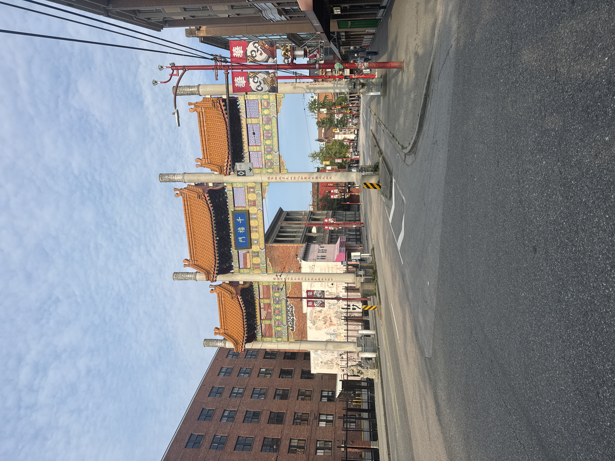 The image captures a street scene in Vancouver's Chinatown, featuring the Millennium Gate. The gate, adorned with traditional Chinese architecture and orange tiled roofs, spans across the road. Beneath the gate, a mural depicting scenes of Chinese life is visible on a building. The street itself is asphalt, with lane markings and a pedestrian walkway. Red lampposts, some decorated with banners featuring pandas and Chinese characters, line the sidewalk. Several buildings, including a tall brick structure and shops with awnings, are visible on either side of the street. The sky is partly cloudy, with a few wisps of white clouds against a pale blue backdrop. There are no people actively engaged in any particular action within the frame, though a few individuals are visible walking on the sidewalk in the distance. The overall impression is of a quiet urban street in the morning.