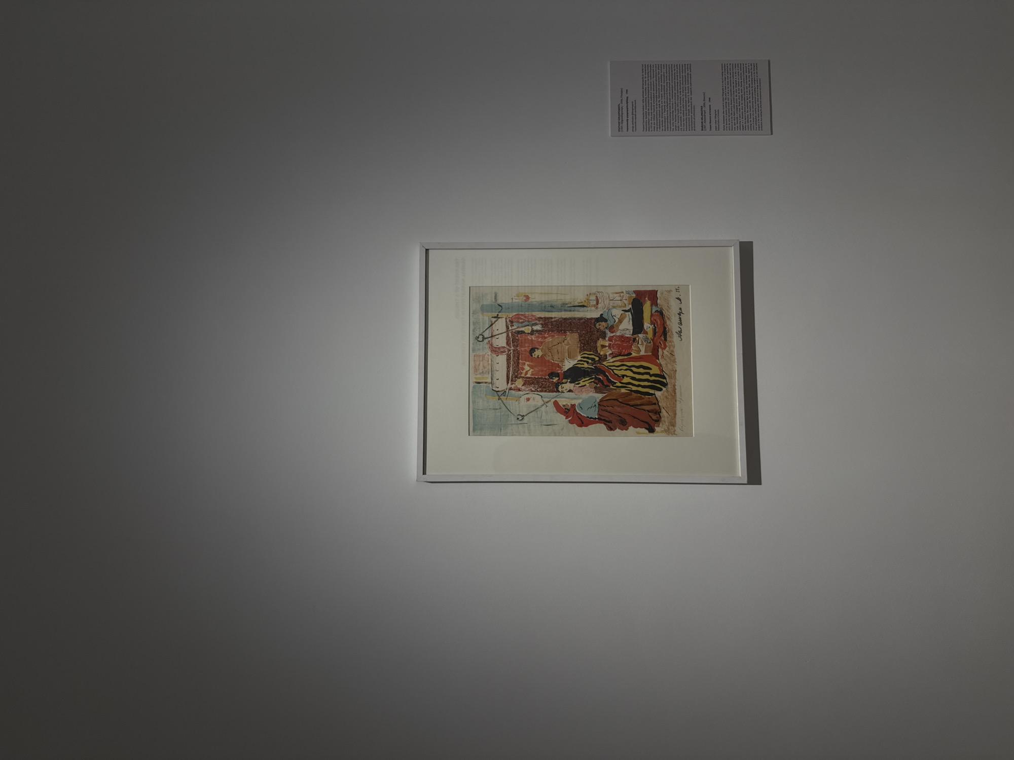 The image depicts an artwork hanging on a gray wall inside a building. The artwork is framed in white and appears to be a print or painting with a scene that features figures, possibly a domestic interior, and color accents including red, yellow, and brown. Above the artwork is a descriptive text, also mounted on the wall, likely providing information about the artwork's title and artist. The building appears to be an art gallery or museum, given the display style. The specific location is Yerevan, Armenia but there are not enough details to know the specific name of the building, gallery, or museum.