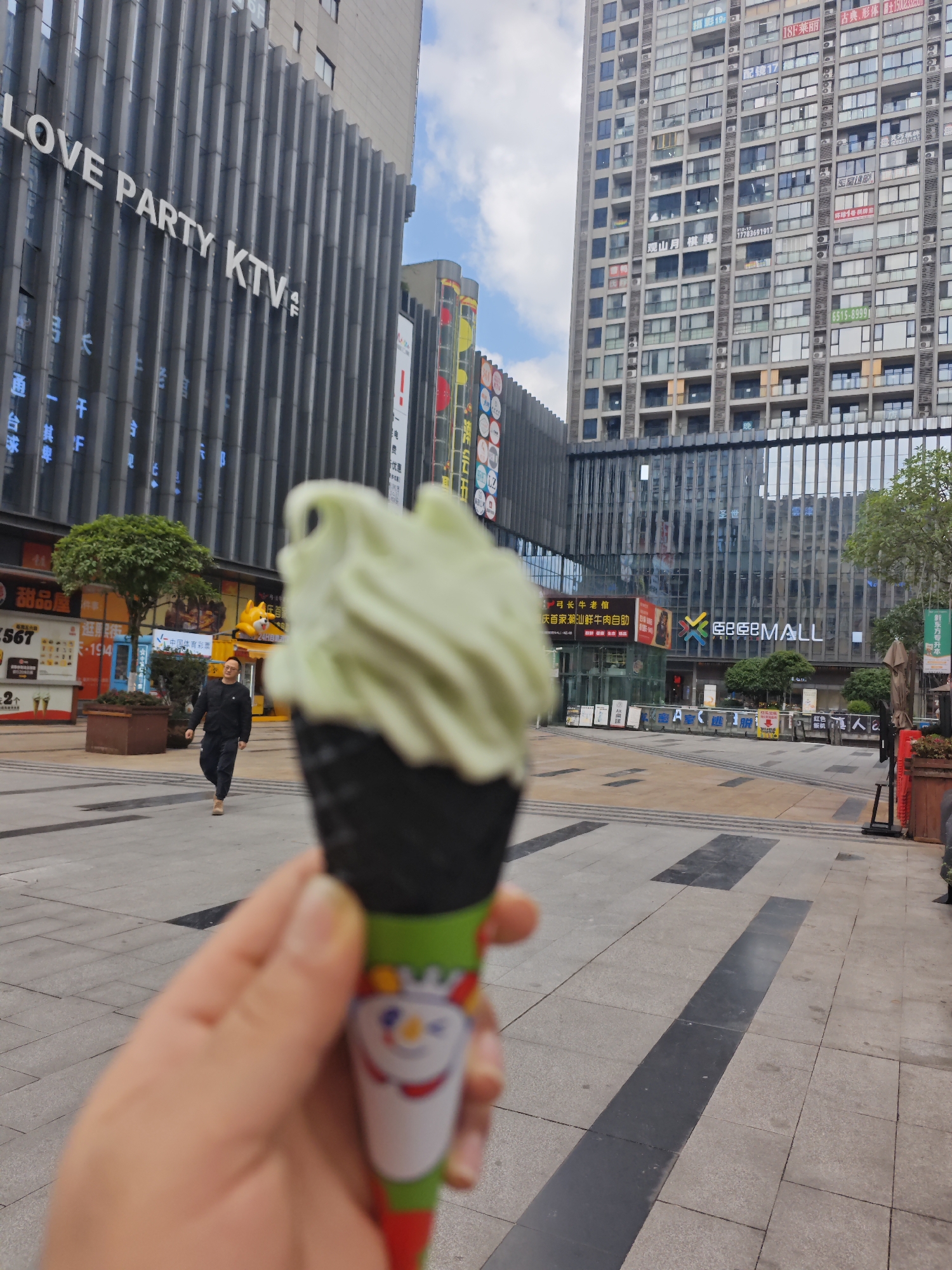 A person holds a pistachio-colored ice cream cone in the foreground. The cone is dark gray with a cartoon character on the holder. The background shows an urban scene in Chongqing High-tech Zone, China. A man in a black jacket and pants is walking left. There is a multi-story building on the left with "LOVE PARTY KTV" written on the facade. Across a paved plaza is another multi-story building with numerous windows and hanging advertisements. In front of that building is "金亞 MALL" in white letters. A few trees and street furniture are visible on the right. The sky is partly cloudy.