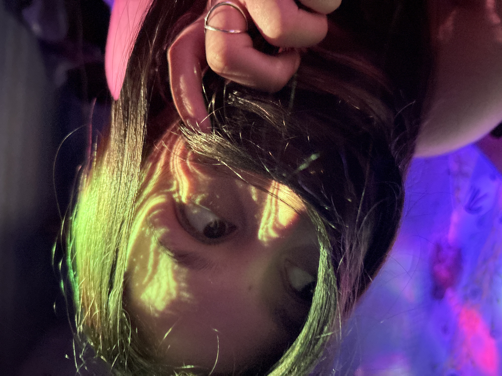 A close-up, upside-down image shows a person's face illuminated with green and yellow patterned light. The person has dark hair, and a hand wearing a silver ring is near the top of the frame. The background has areas of purple and darker shades. The location is not discernible from the image.
