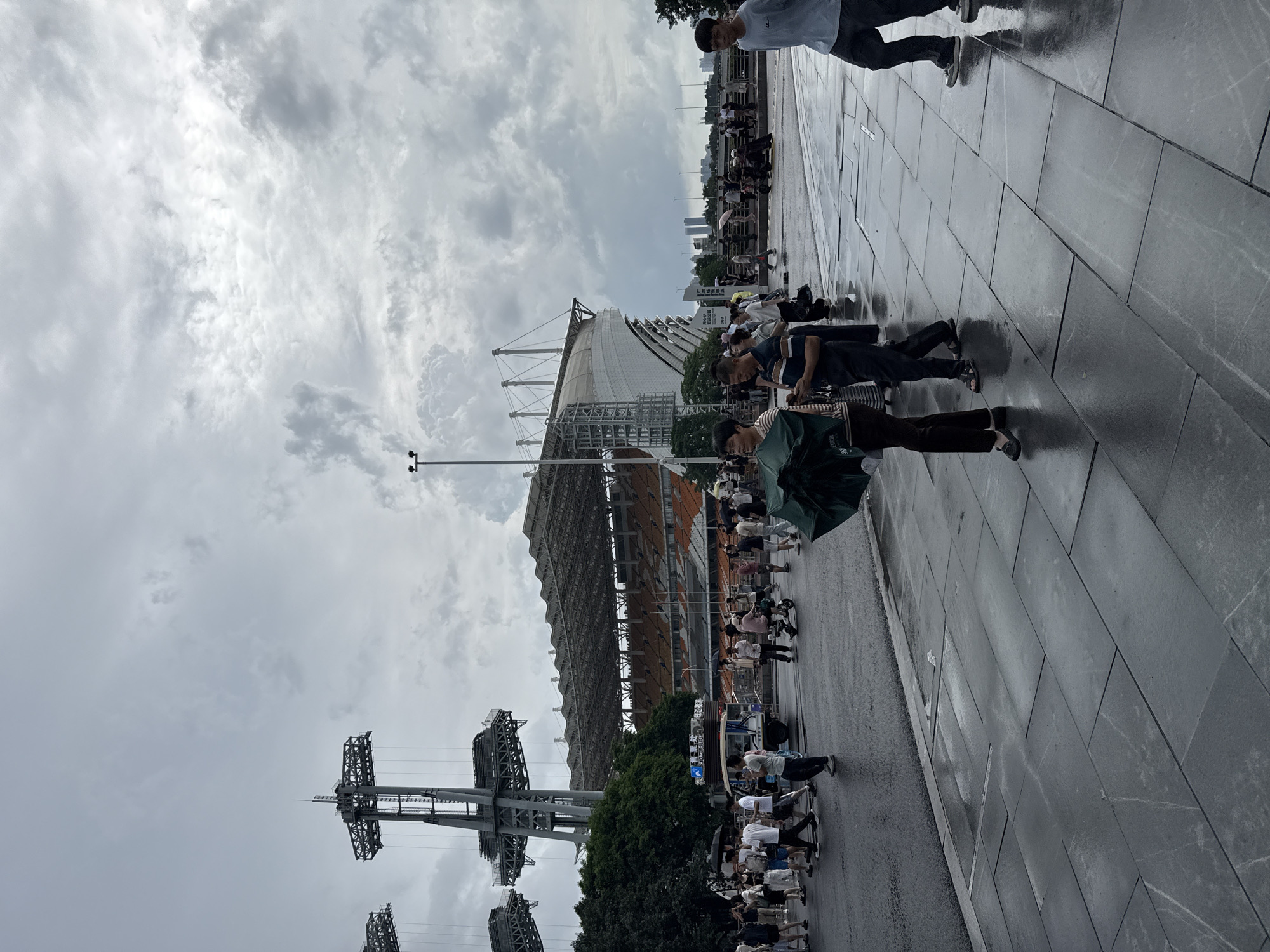 This is a street-level, eye-level, medium shot of a public area with a stadium in the background. The sky is overcast and filled with clouds. In the foreground, a wet, paved walkway with large, rectangular tiles reflects the light. Several people are walking on the walkway and the adjacent street.
To the left, a woman holding a green umbrella walks towards the viewer. She is wearing dark pants and a lighter-colored top. Behind her, a man in a striped shirt and dark pants is looking at his phone. Further left, a group of people are walking in various directions. One individual is pushing a small cart.
In the middle ground, a stadium with orange seating is visible behind a row of trees. Two large, metal structures with a complex, lattice-like design stand in front of the stadium. People are gathered in front of the stadium and near the structures. A light-colored, three-wheeled vehicle is also present in this area.
In the background, the skyline of a city is visible, with several tall buildings. The overall impression is that of a busy public space, possibly a tourist attraction, on a cloudy day, with evidence of recent rain on the ground. The scene is located in Tianhe District, China, likely near the Tianhe Stadium given the prominent structure in the background.