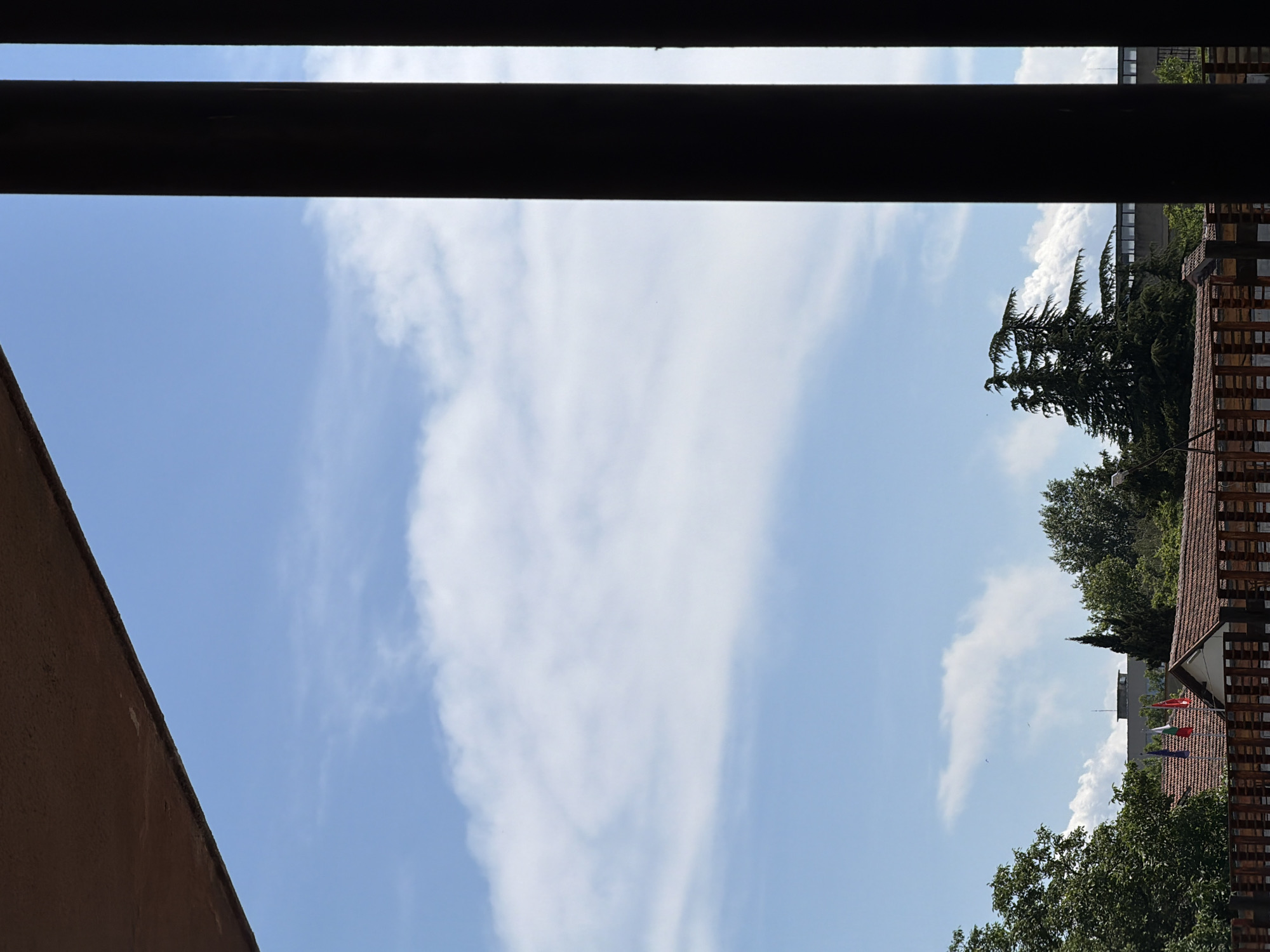 The image captures a view through dark vertical beams and a brown overhang, framing a bright blue sky with scattered white clouds. In the middle ground, a cluster of green trees, including a tall, feathery pine, stands in front of a building with a dark, horizontal band. Below the trees, a red-tiled roof is visible, with a wooden fence in the foreground. Three flags, one red, one white and green, and another red, are mounted on poles above the roof. The scene appears to be an outdoor setting in a city, possibly on a sunny day.