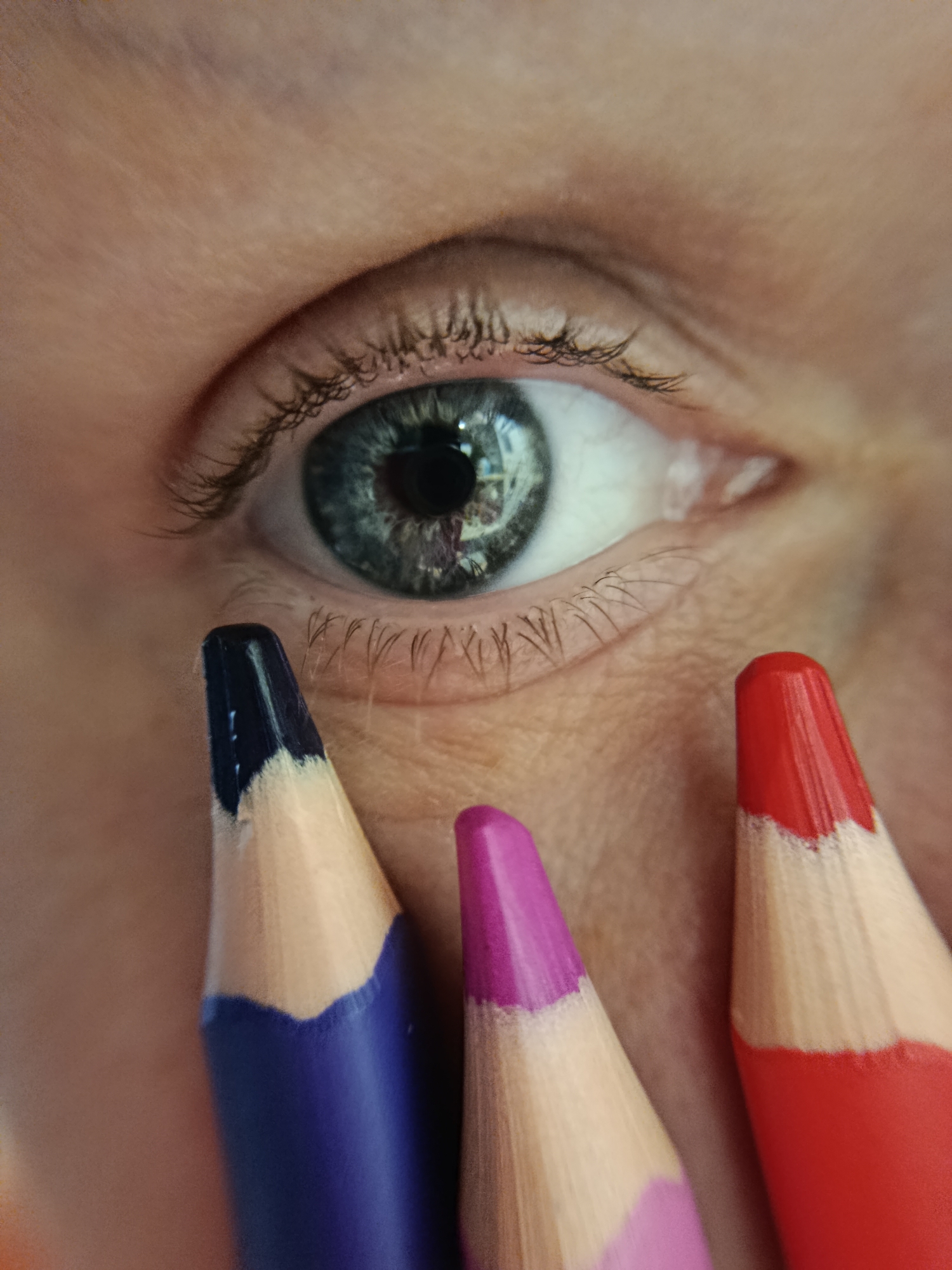 The image is a close-up shot of a person's eye with three colored pencils in the foreground. The eye is open and appears to be blue-green. The pencils are positioned such that their colored tips are pointing towards the eye. From left to right, the pencils are dark blue, purple, and red. The skin around the eye is visible, with some fine lines and eyelashes. The background is out of focus, showing the person's skin. There is no indication of the city of Фоки, Russia, or any specific environment.