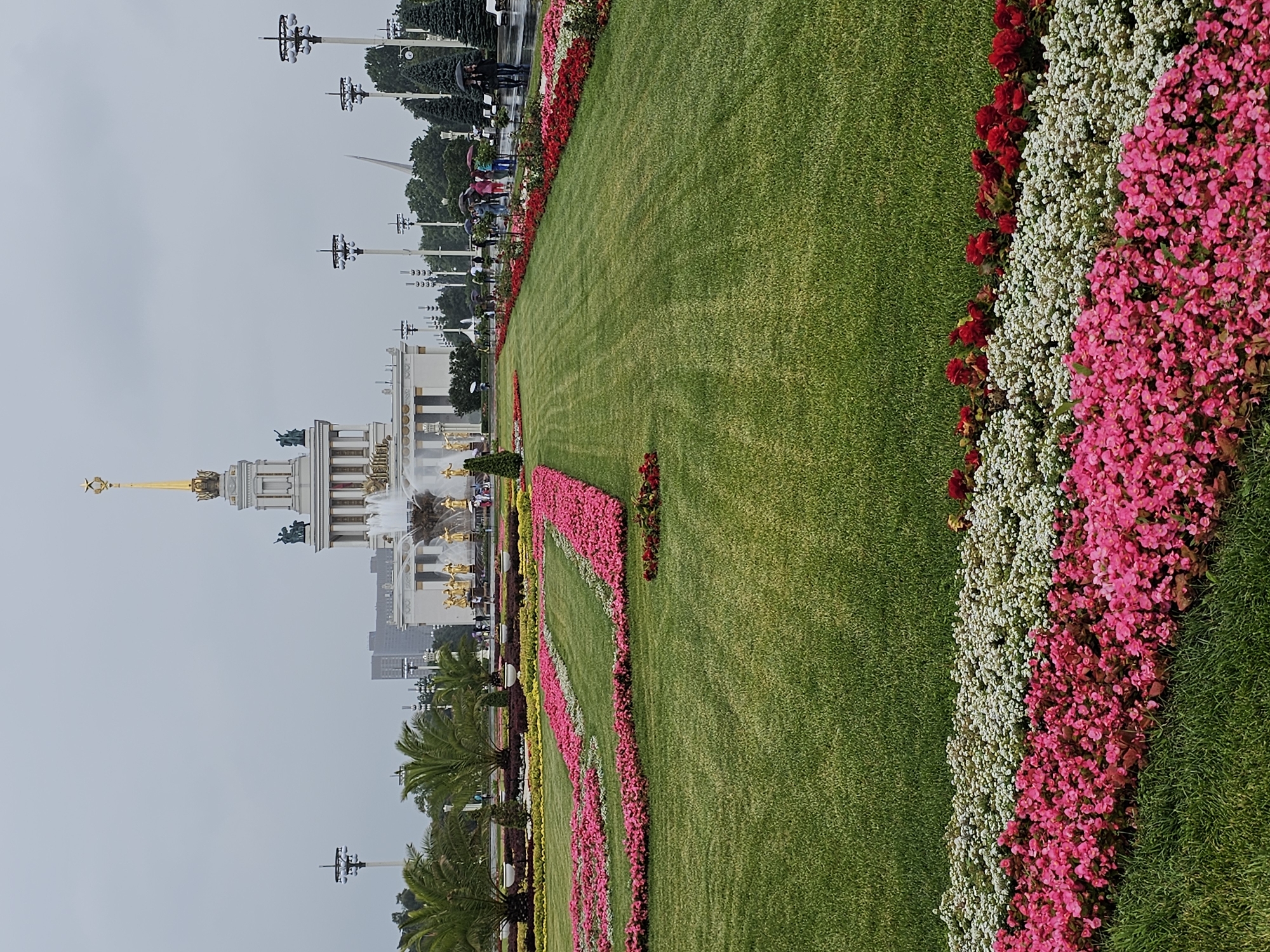 The image shows a frontal view of the "Friendship of Peoples" fountain at VDNKh (Exhibition of Achievements of National Economy) in Moscow, Russia. The fountain is adorned with golden sculptures of Soviet republics. In the background, the main pavilion of VDNKh is visible, a grand Stalinist Empire style building topped with a golden spire. Lush green lawns and meticulously arranged flower beds in vibrant shades of pink, white, and red fill the foreground and midground, leading towards the fountain and pavilion. Several people are walking around with umbrellas, suggesting it is a cloudy or rainy day. Lampposts line the pathways, and palm trees add a touch of tropical flair to the landscape.