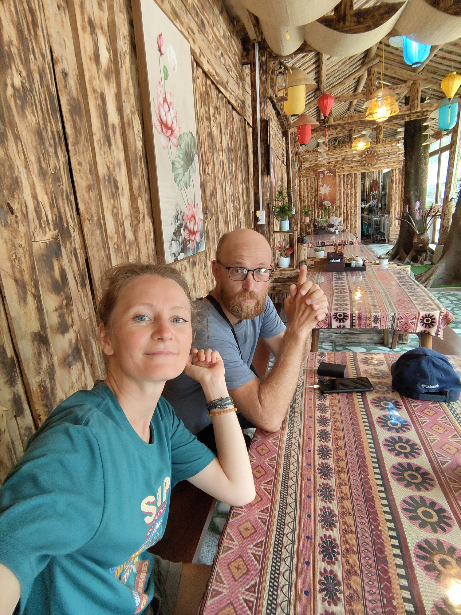 A waist-up shot captures a woman and a man seated at a table in what appears to be a restaurant or cafe in Thành phố Hà Giang, Vietnam. The woman, positioned on the left, wears a teal t-shirt and rests her head on her hand. She has fair skin and blue eyes, and smiles at the camera. To her right, the man, with a bald head and a beard, wears glasses and a gray t-shirt, points a finger upwards and looks toward the camera. The table between them is covered with a patterned cloth, and there is a phone, a battery charger, and a blue baseball cap visible on the table. The establishment's interior features wooden walls adorned with a floral painting, and there are colorful lanterns hanging from the ceiling. The overall atmosphere suggests a casual and rustic setting.