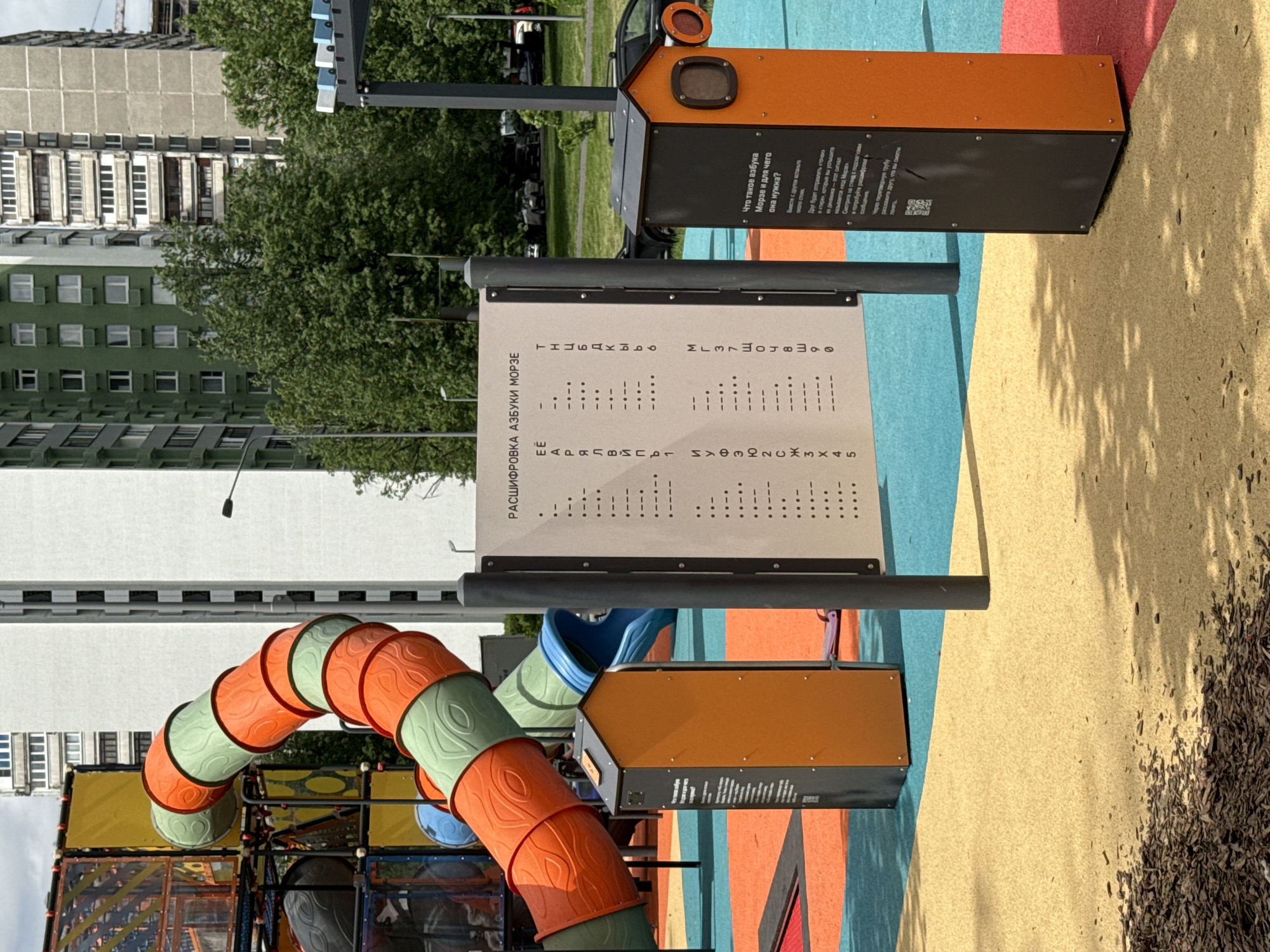 The image shows a playground in Moscow, Russia. In the center of the frame, there's a sign displaying the Morse code alphabet in Russian, labeled "Deciphering the Morse Alphabet". The playground features a bright color scheme, with orange, blue, yellow, and red sections. In the background, a multi-story residential building is visible, with green trees obscuring parts of it. The playground equipment includes an orange and green tunnel slide, part of a larger climbing structure. The photograph is taken at a slightly angled perspective.